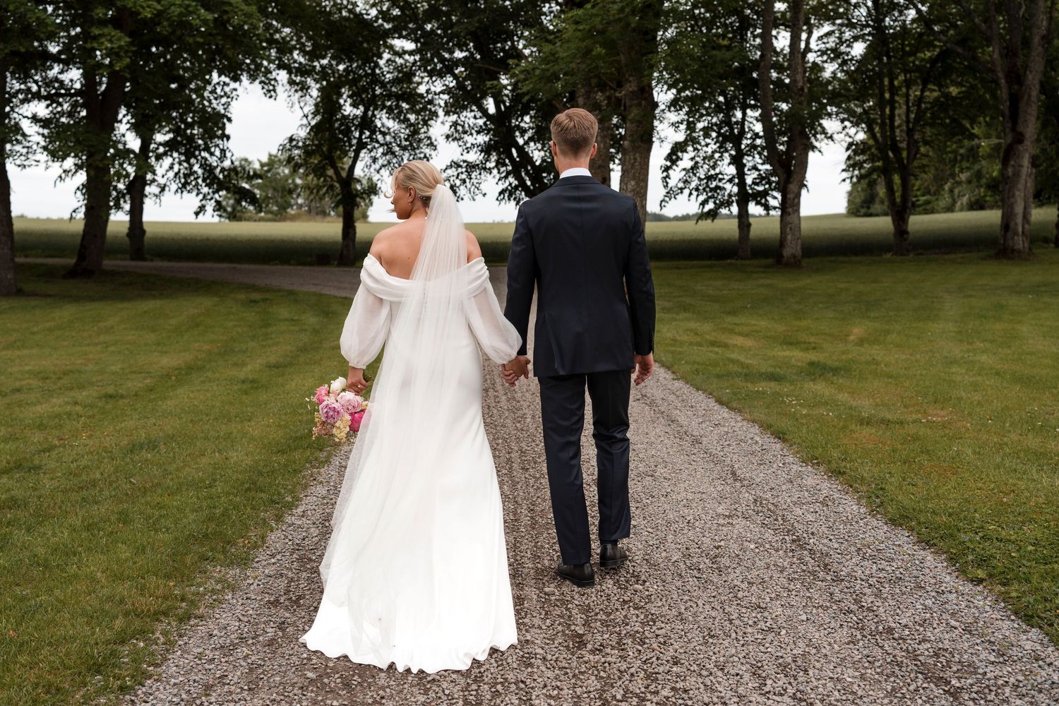 A couple in wedding attire walks down a gravel path surrounded by green grass and trees.