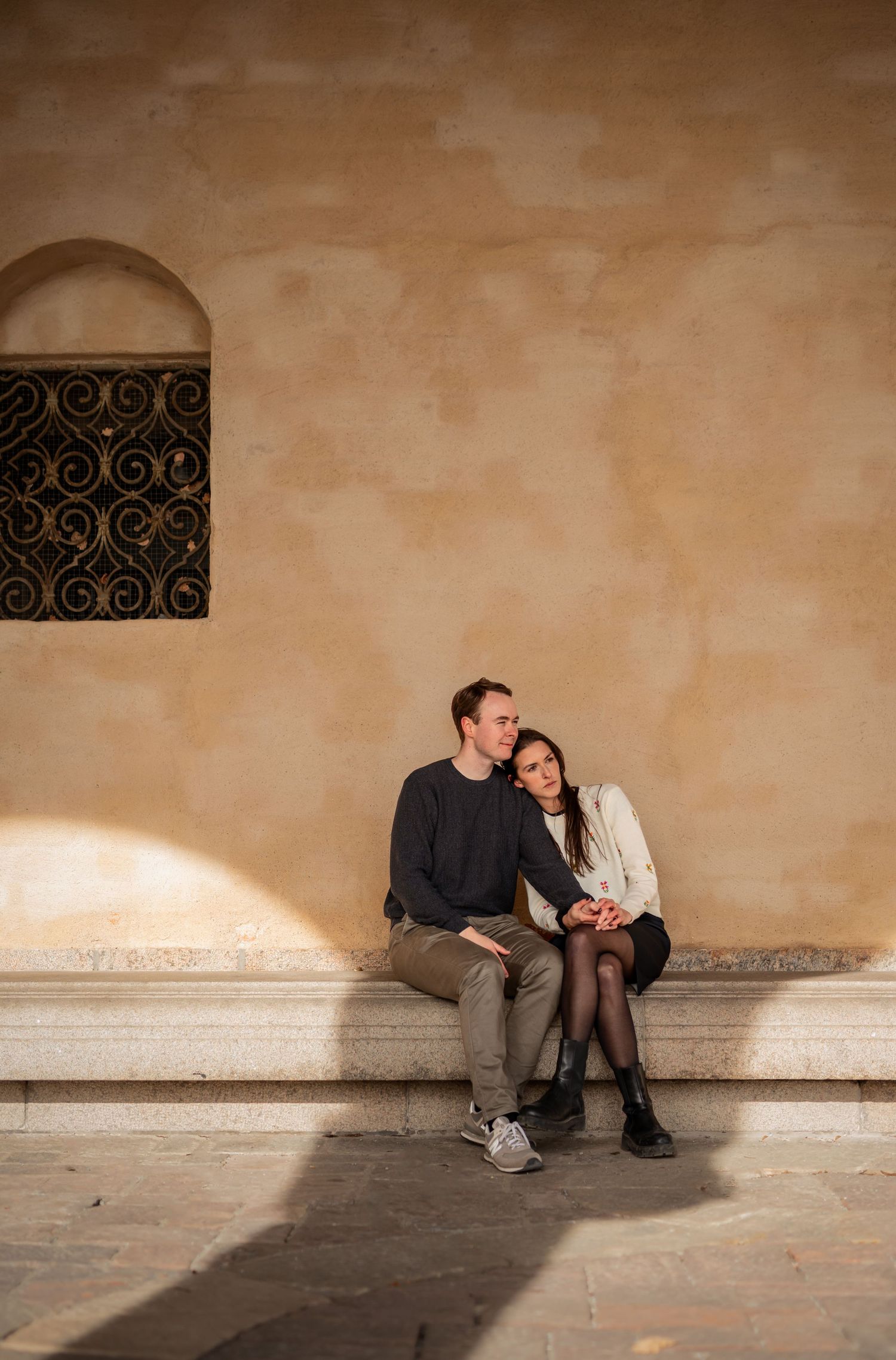 A couple sits together on a stone bench against a beige wall with decorative ironwork window in evening light.