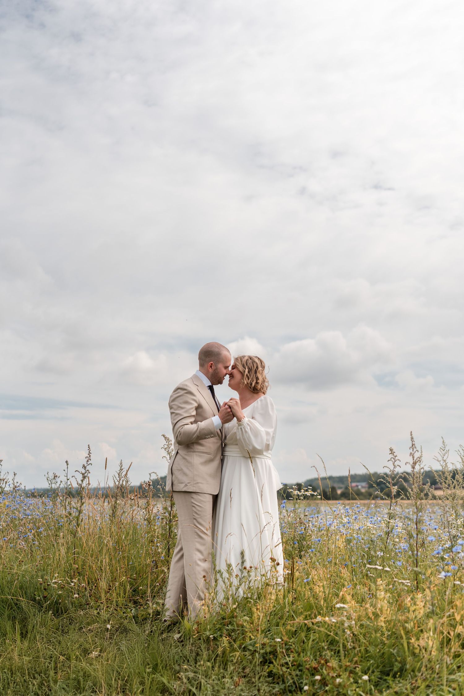 A couple in wedding attire embrace on a grassy hillside overlooking a lake on an overcast summer day.