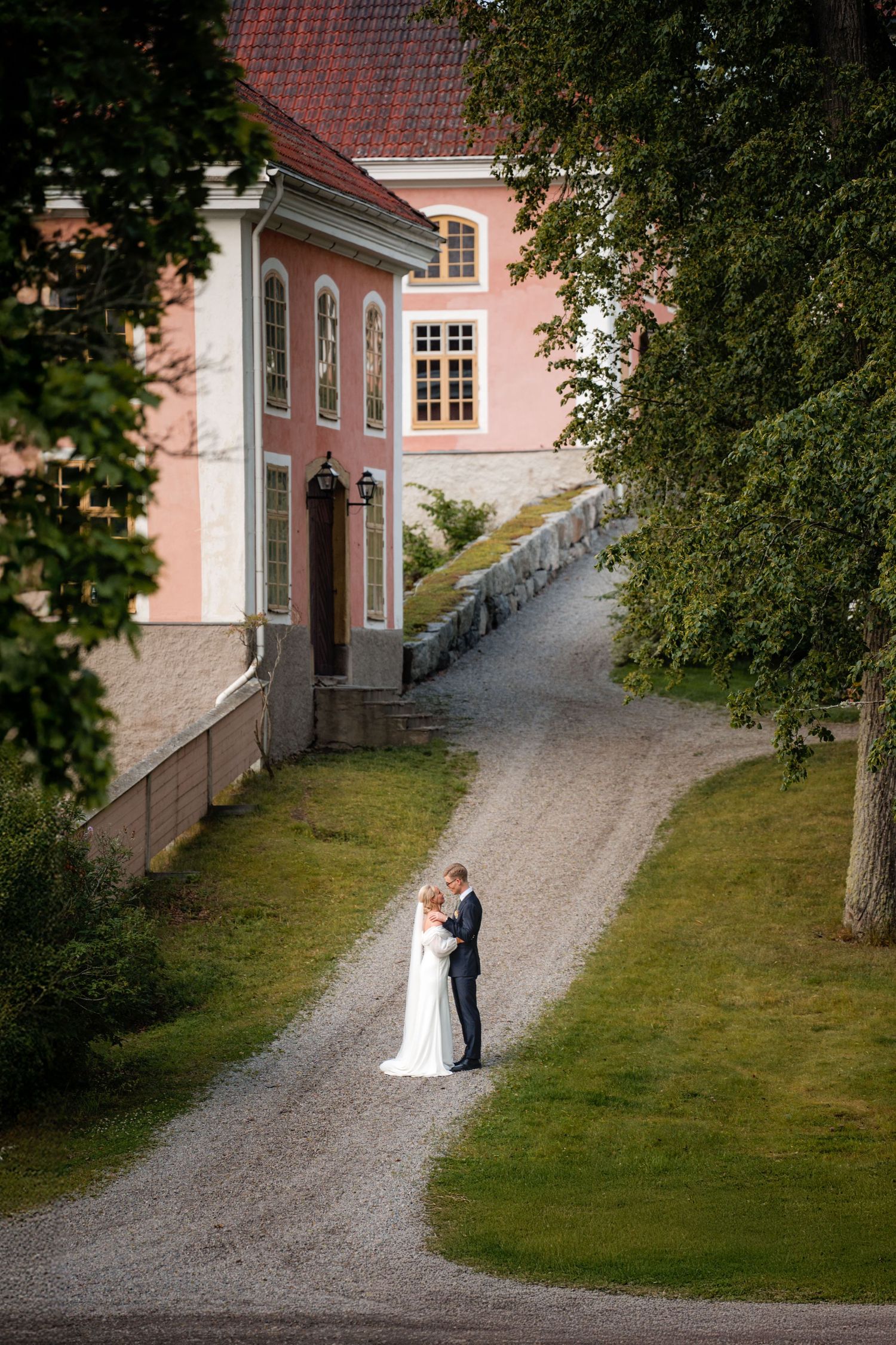 A couple in wedding attire walks along a winding gravel path beside a pink historic building on their special day.