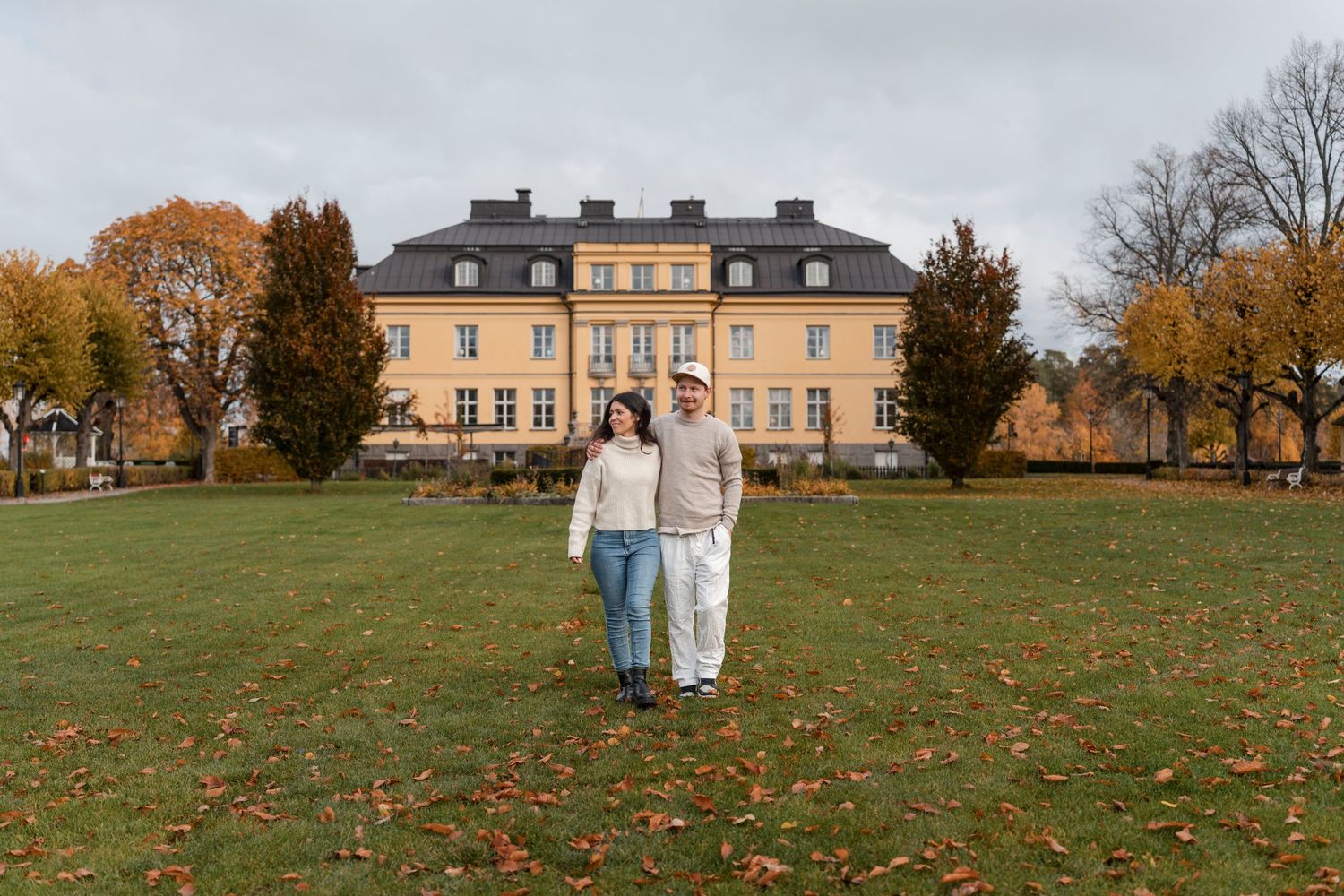 Elegant manor house with yellow facade surrounded by autumn trees and sprawling green lawn on a fall day.
