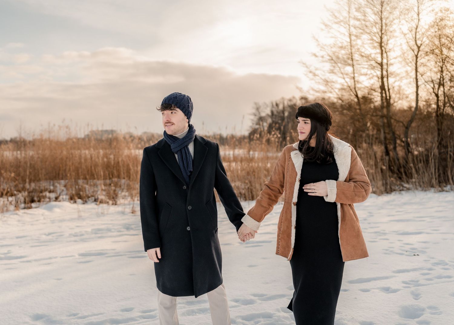 Couple holding hands walking in snowy winter landscape at sunset, man in black coat and woman in brown jacket.