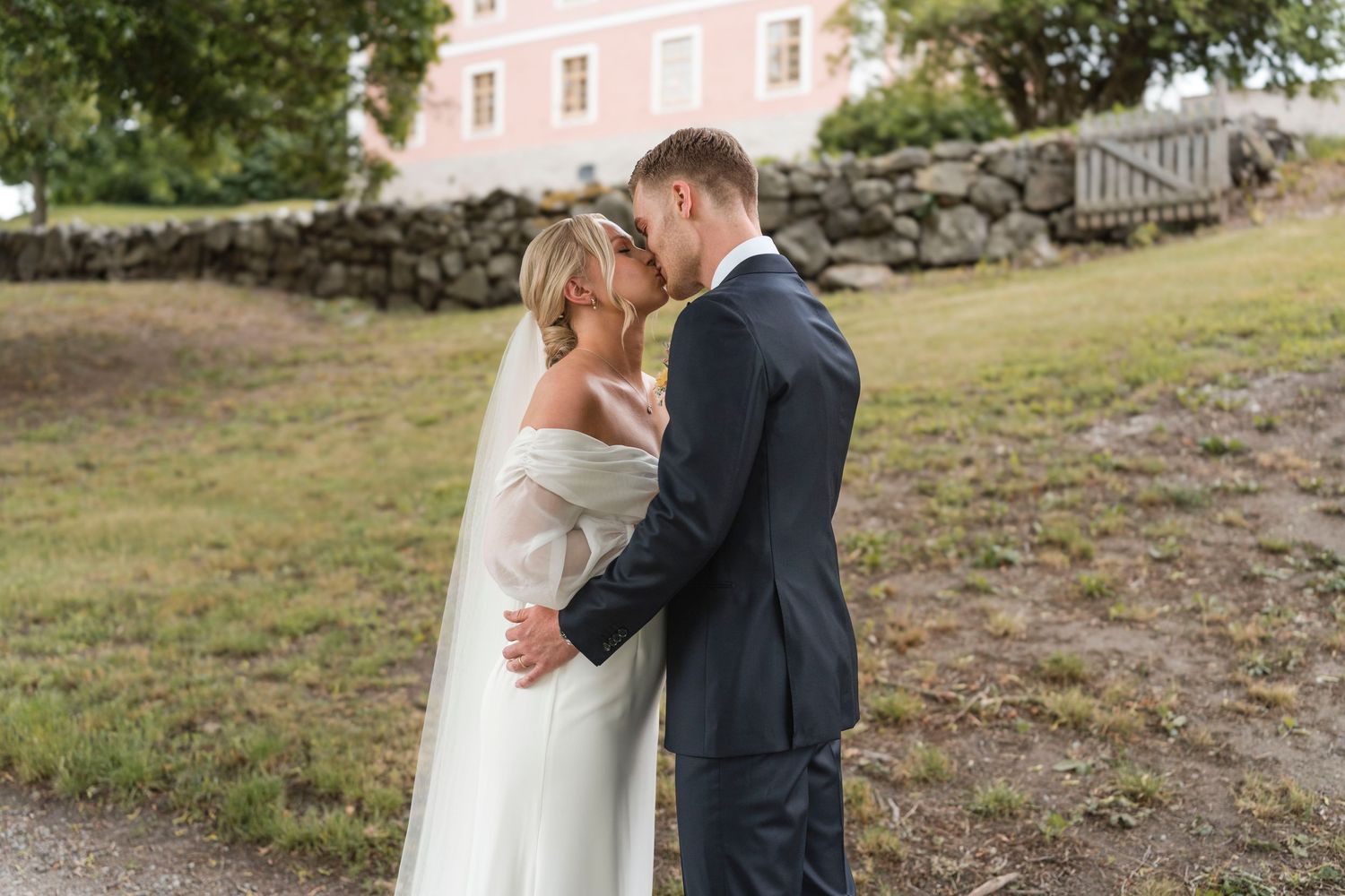 A couple shares a romantic kiss in their wedding attire while standing in a scenic outdoor setting.