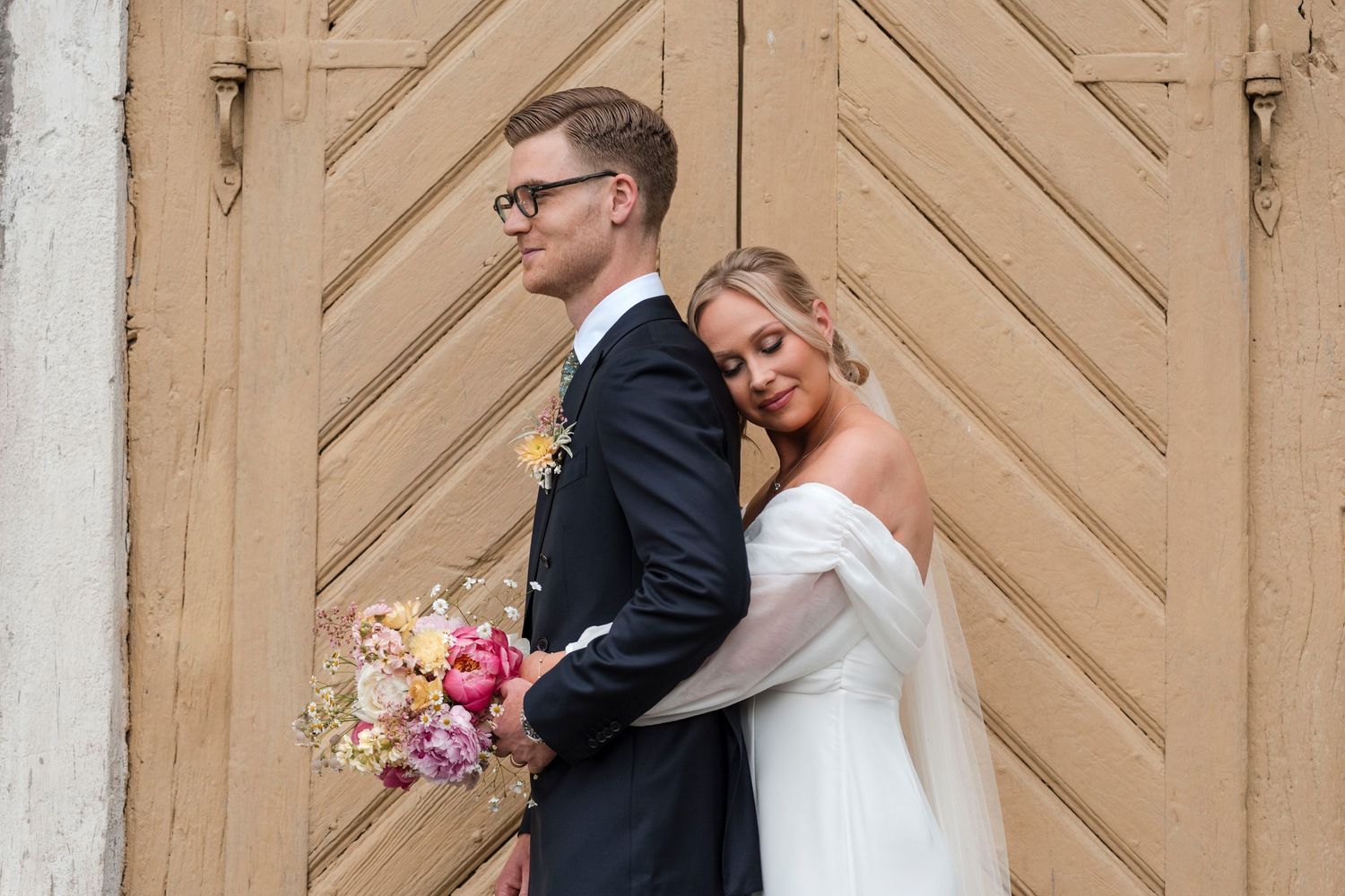A couple shares a tender moment in front of a beige chevron patterned door while holding a pink floral bouquet.