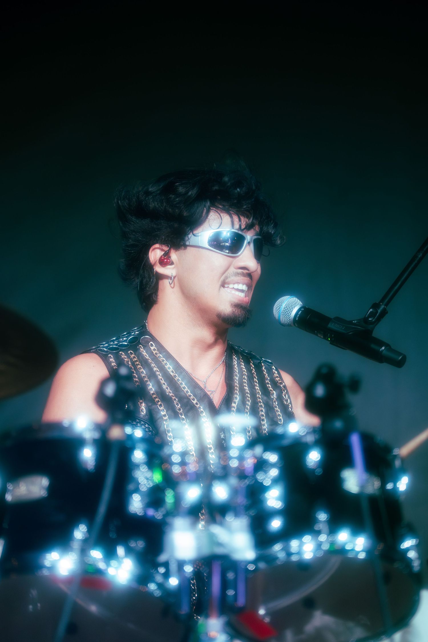Male rock drummer performing on stage wearing sunglasses and studded vest with microphone nearby.