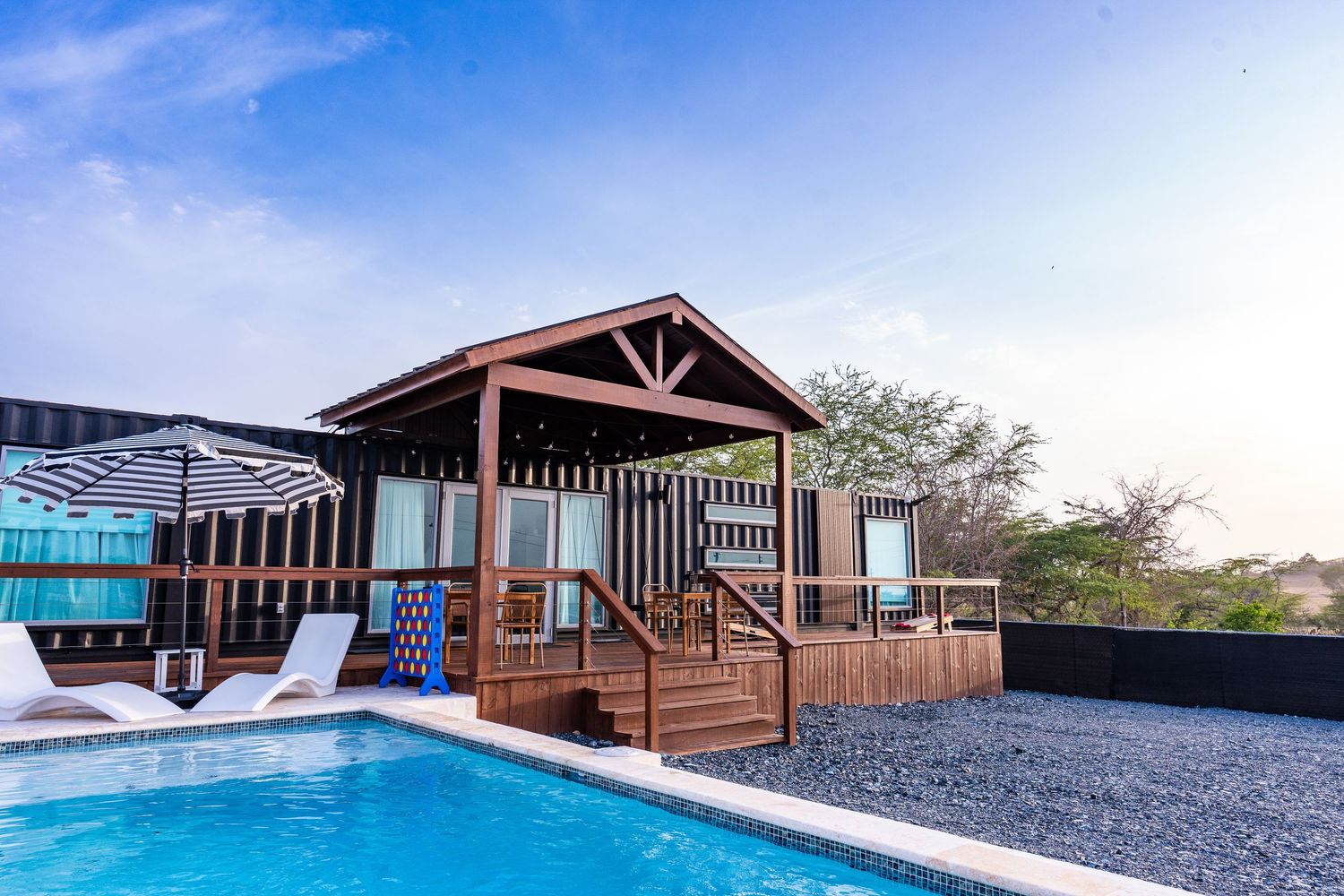 Wooden bungalow with covered deck beside a swimming pool under a clear blue sky.