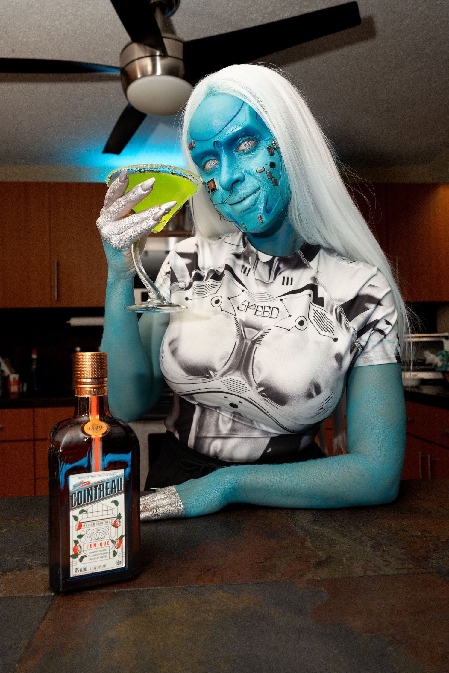 Blue robot makeup costumed person smiling while holding green cocktail with Cointreau bottle.