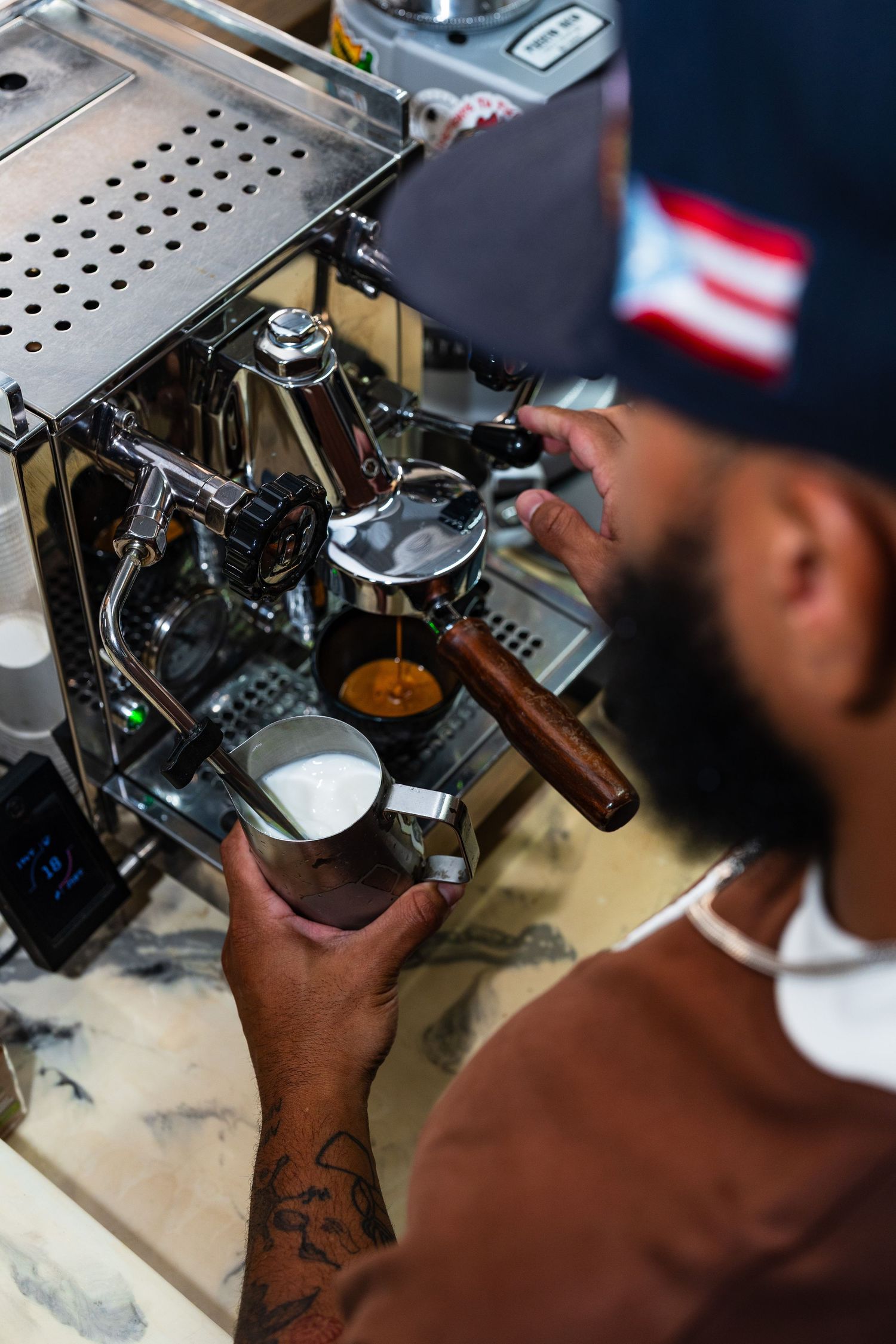 Bearded barista wearing a cap extracting espresso on a professional machine while steaming milk nearby.