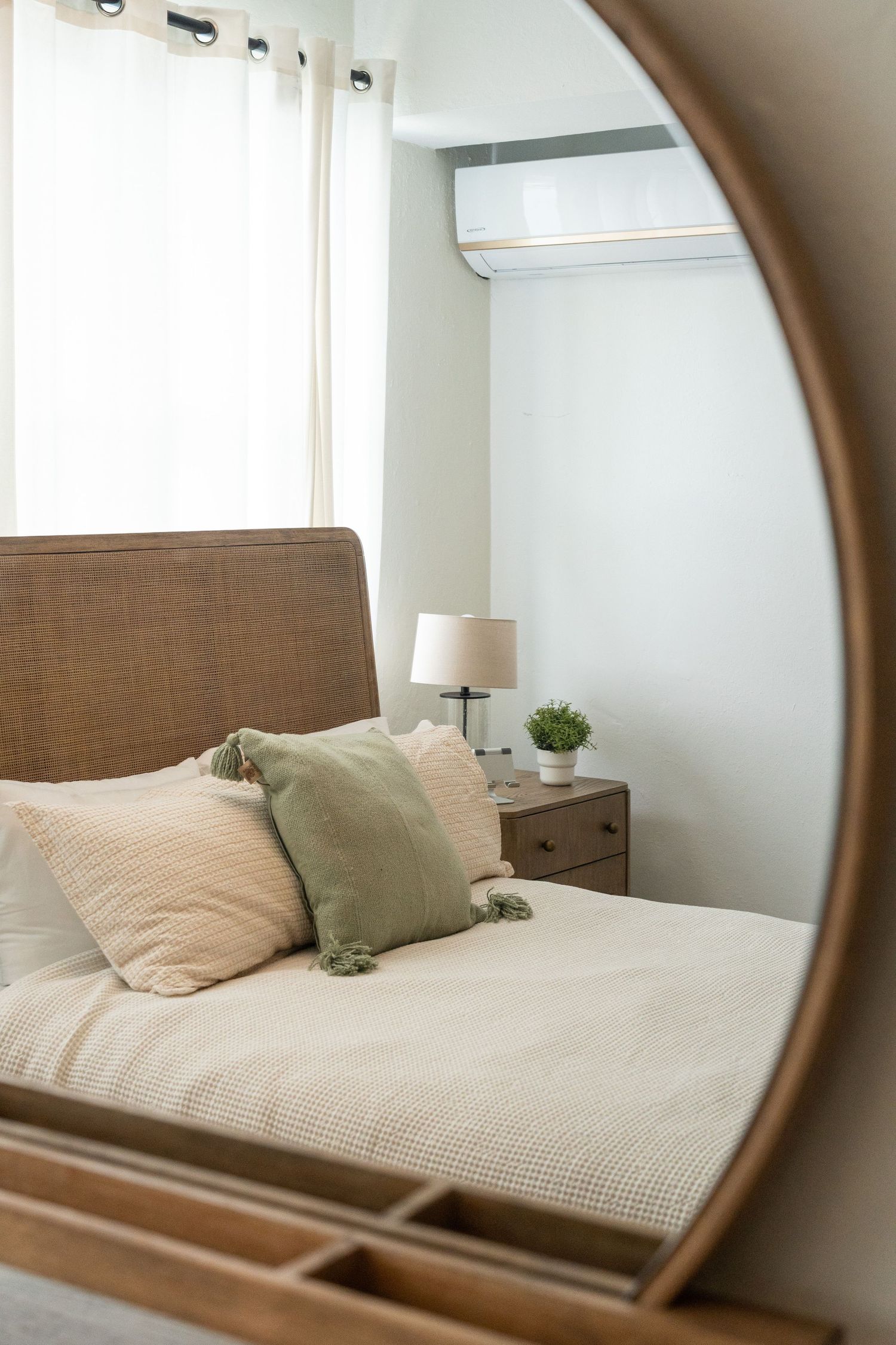 Cozy bedroom reflected in oval wooden mirror with green pillows and neutral bedding.