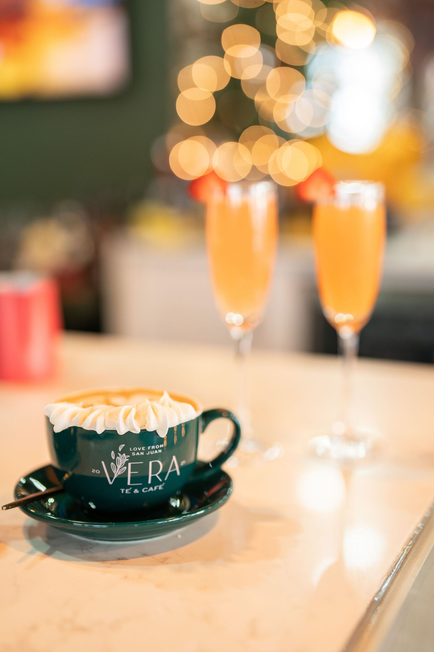 Green Vera Te and Cafe branded cappuccino with whipped cream beside two champagne flute cocktails.