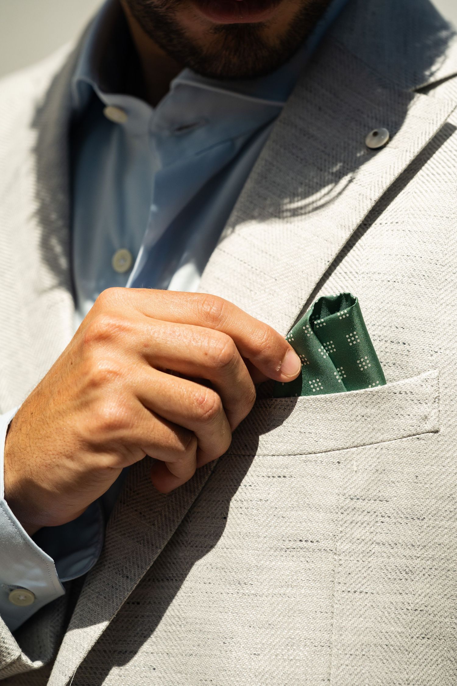 Close-up of mans hand adjusting green pocket square in beige blazer pocket.