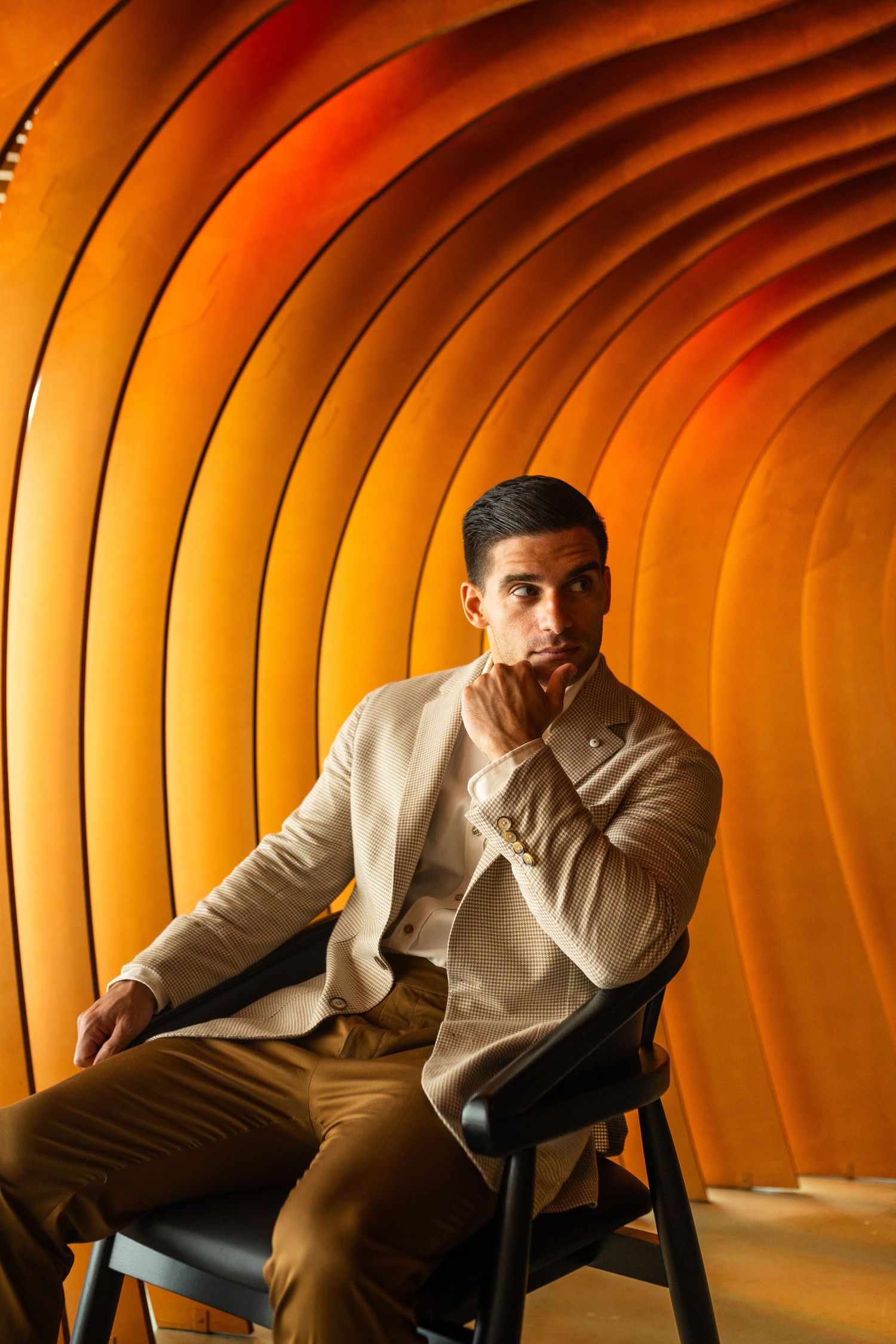 Stylish man in cream blazer and tan pants posing thoughtfully against glowing orange arch backdrop.