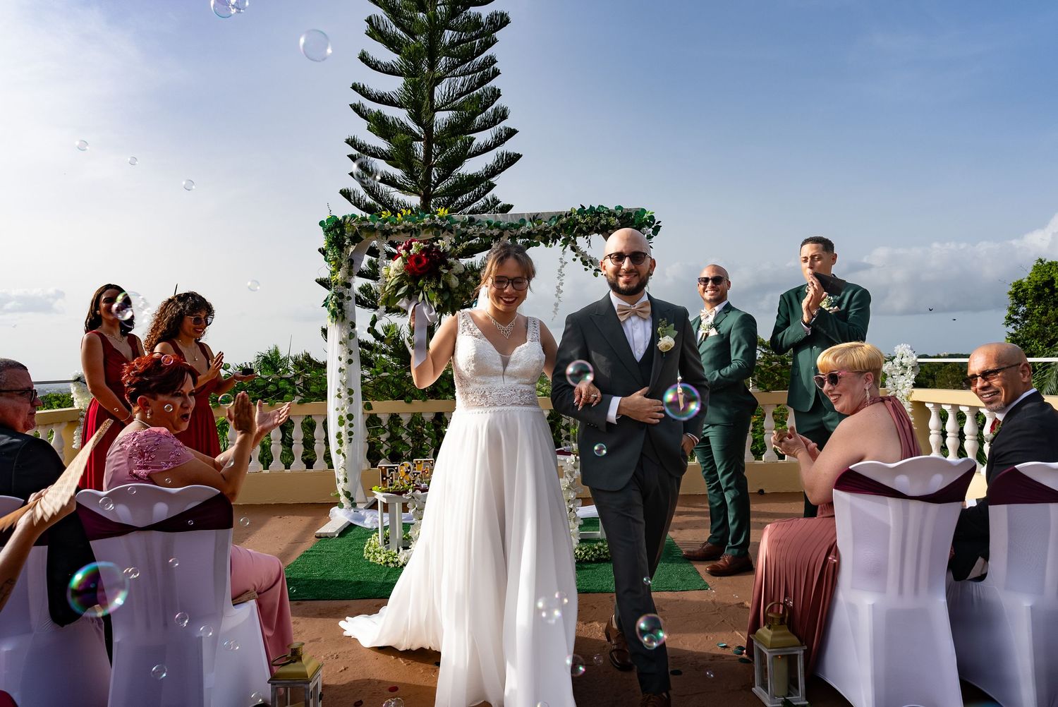 Joyful newlywed couple walking down outdoor wedding aisle surrounded by bubbles and guests at scenic venue.