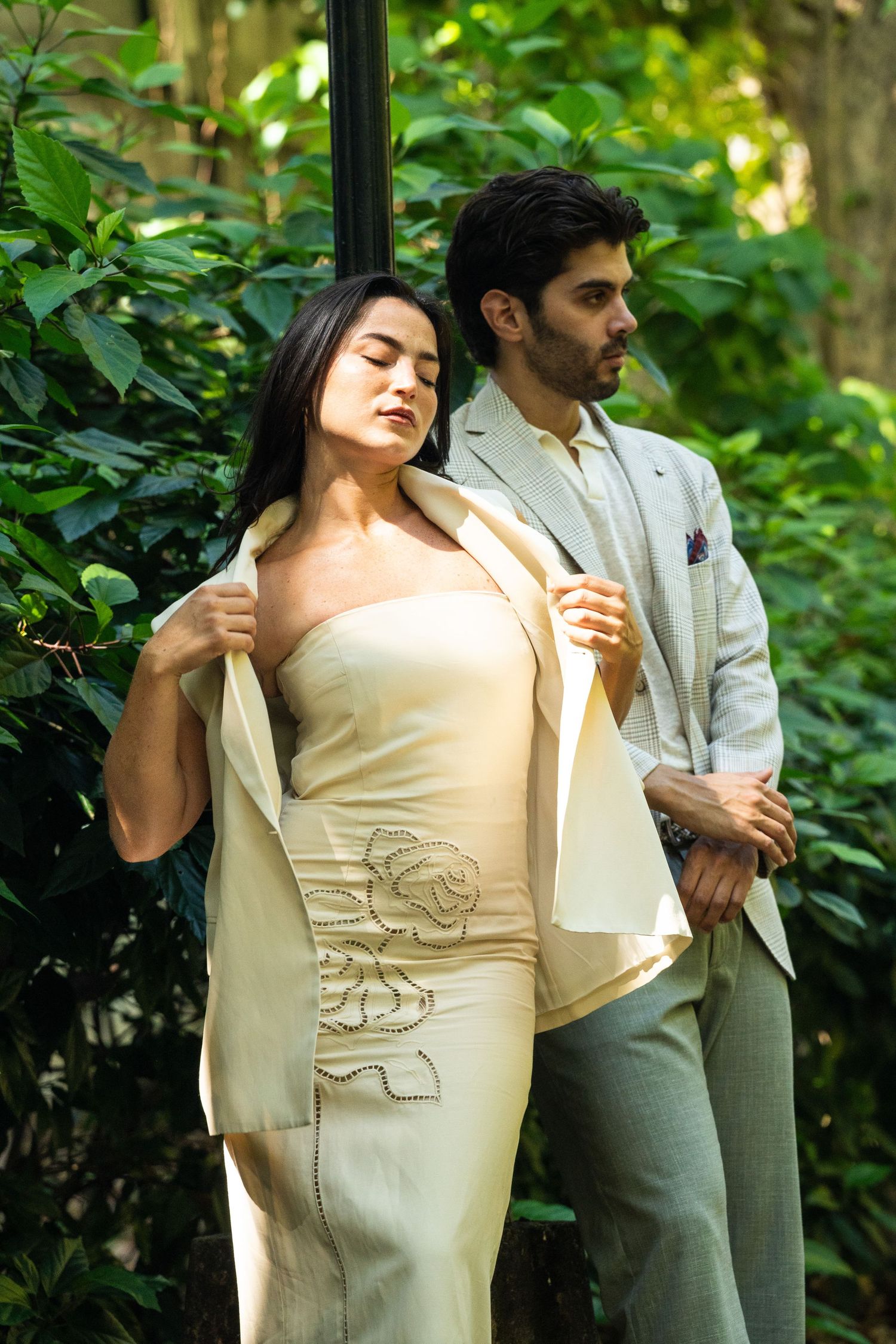 Elegant couple posing outdoors, woman in cream strapless gown and man in light gray suit among lush green foliage.