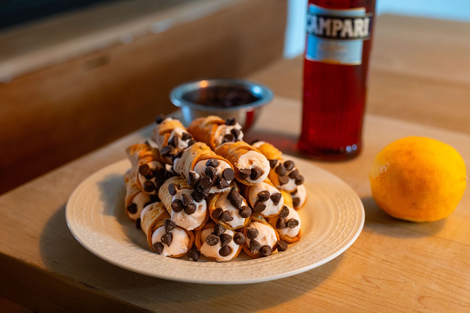 Plate of mini pastries with chocolate chips beside Campari bottle and orange on wooden table.