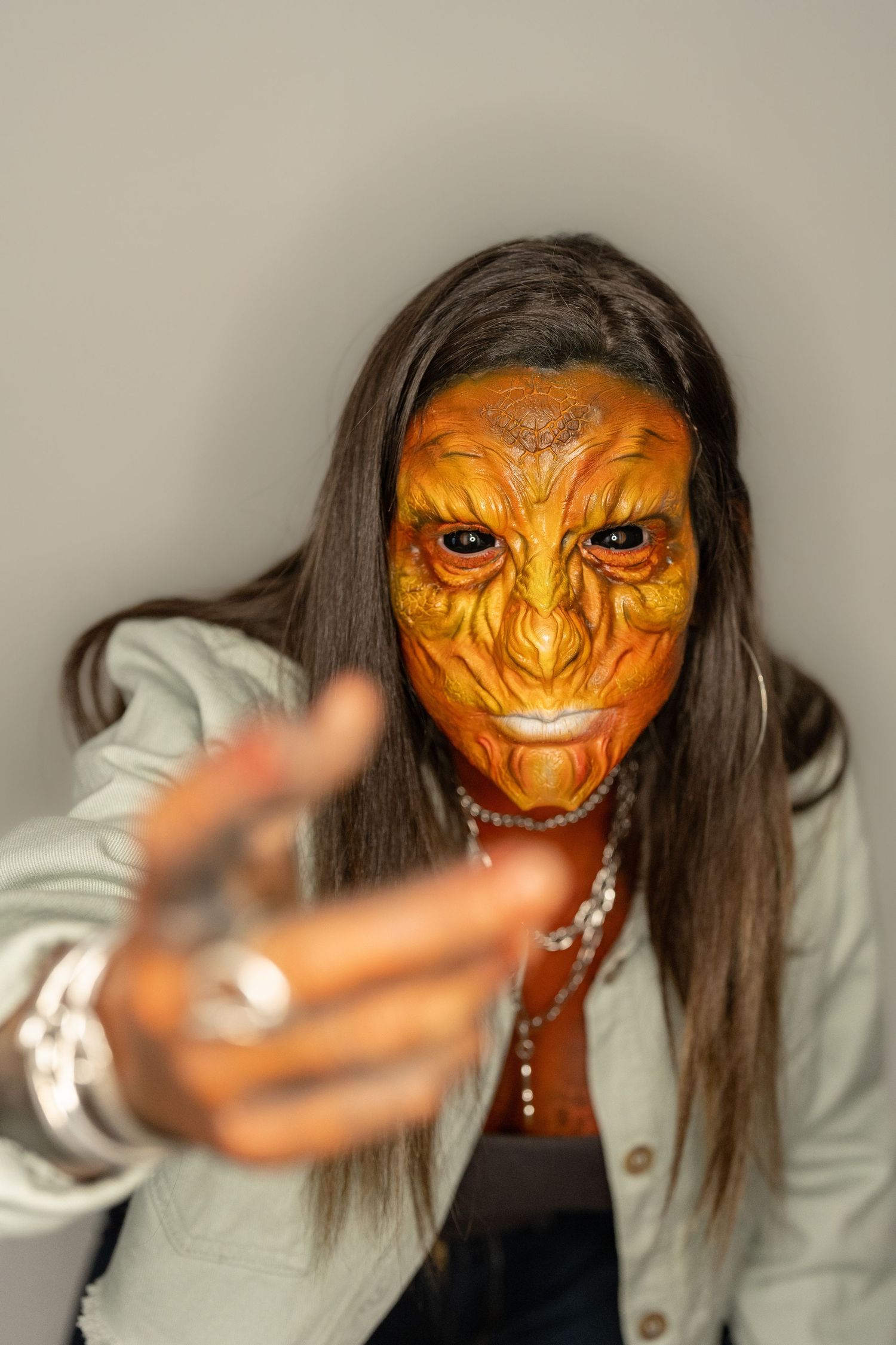 Person wearing orange reptile mask in denim jacket reaches toward camera against gray background.