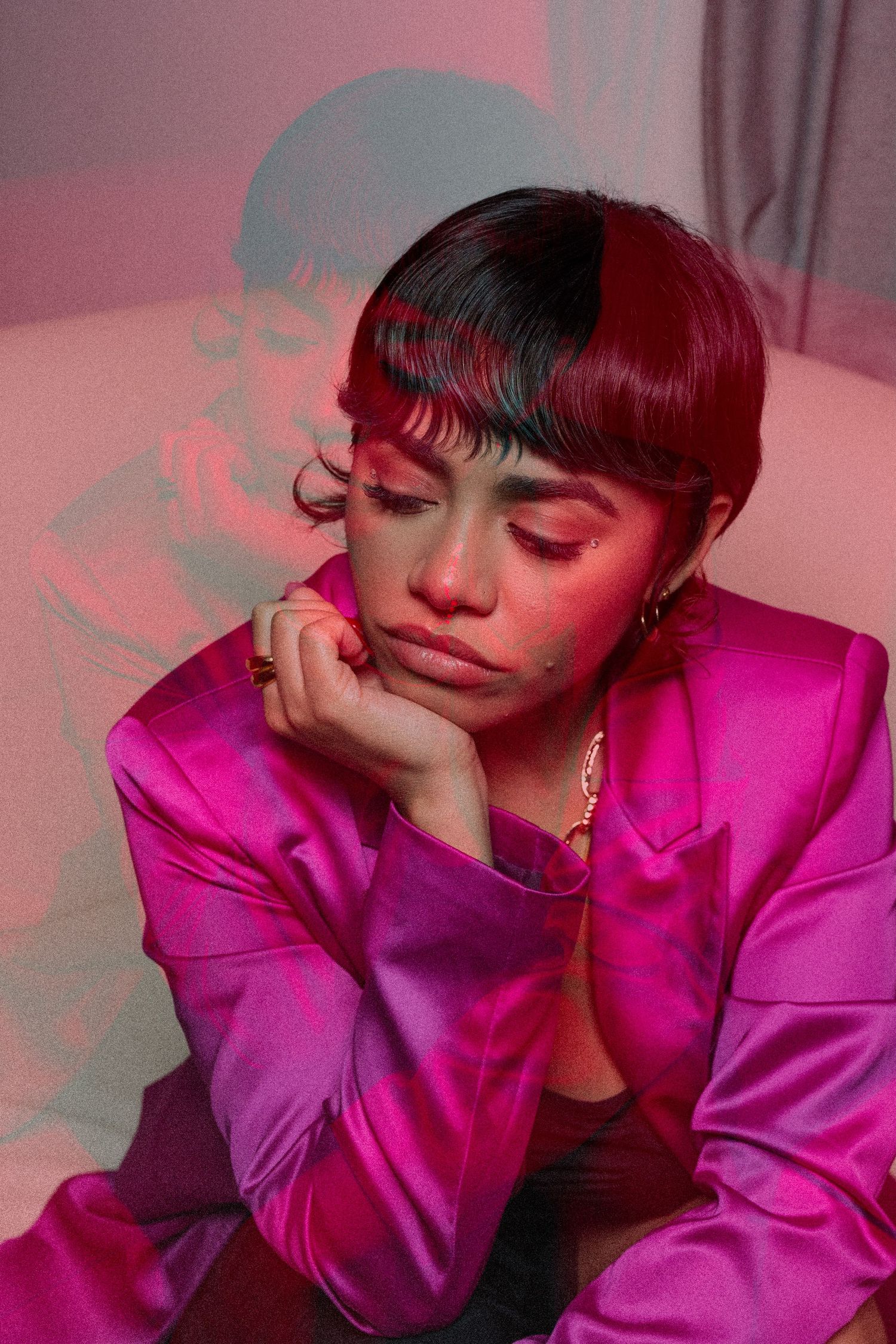 Young woman in pink satin blazer with short dark hair poses thoughtfully under pink and blue lighting.