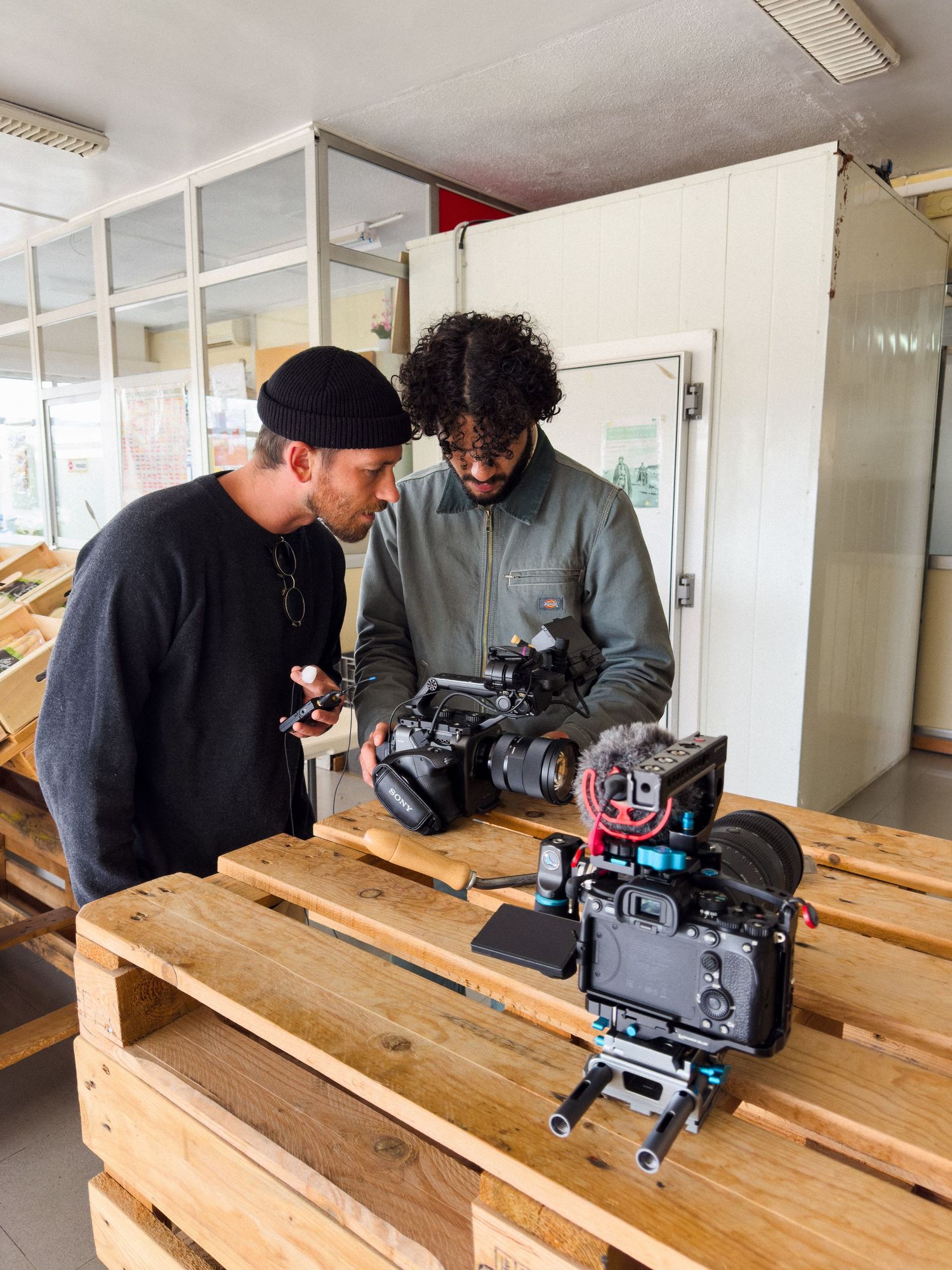 Two filmmakers review camera equipment on wooden pallets in an indoor studio setting.