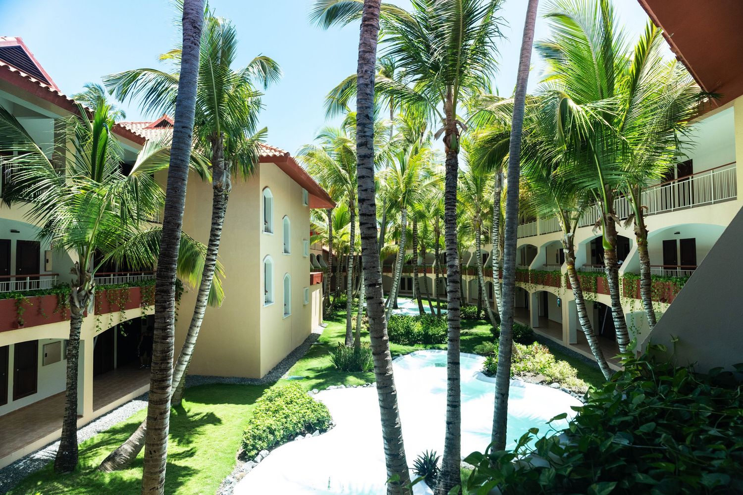 Tropical resort courtyard with tall palm trees, lush greenery, and colorful Spanish-style buildings.