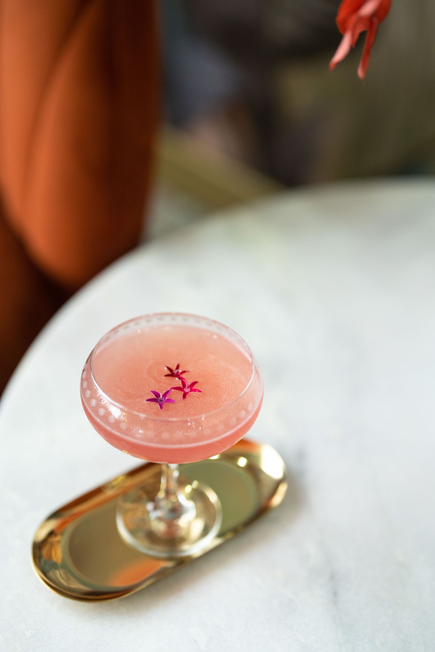 Pink cocktail in coupe glass garnished with purple flowers on gold tray on marble table.