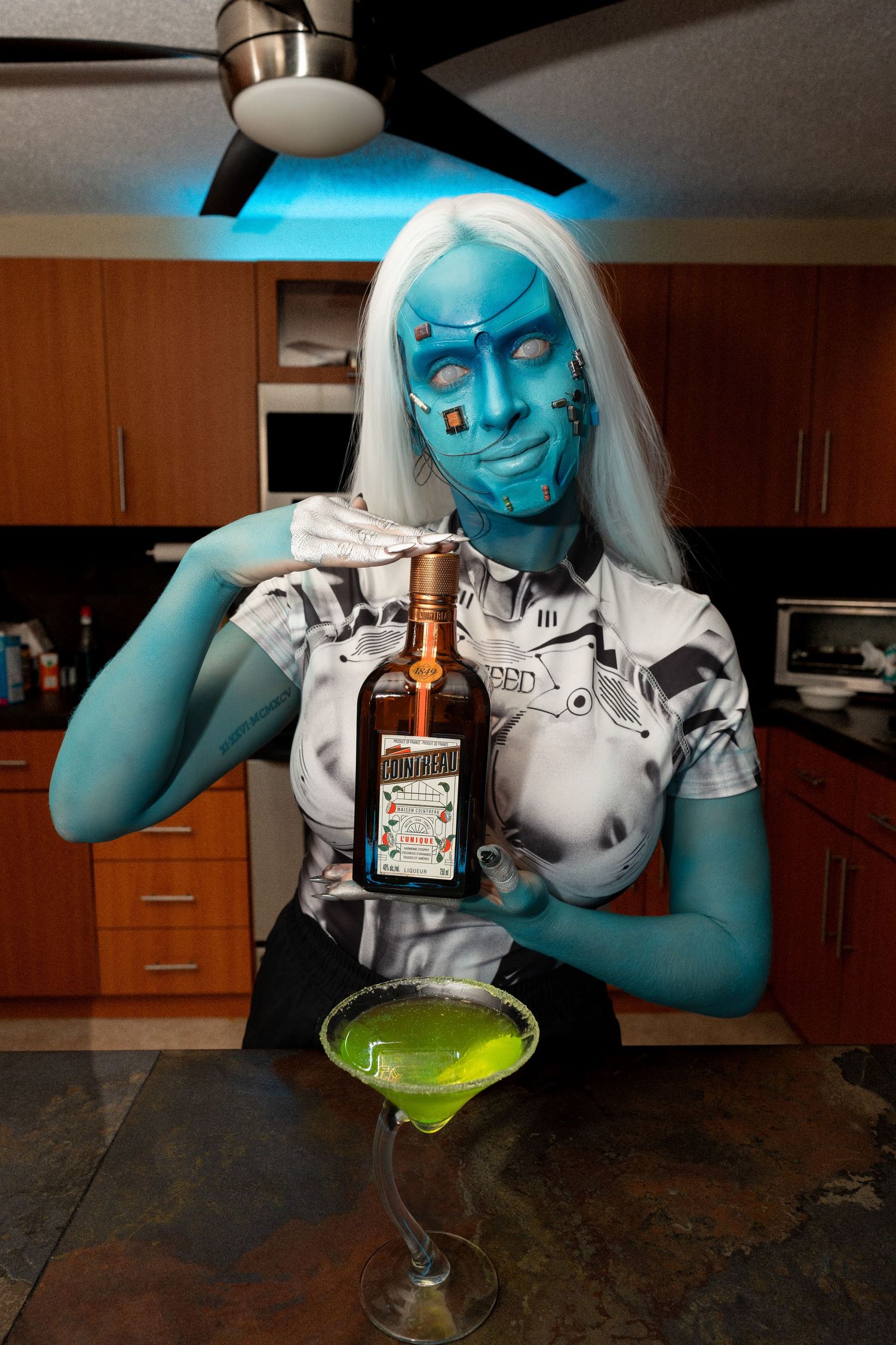 Blue android costumed person pouring Cointreau into large green martini glass in kitchen.