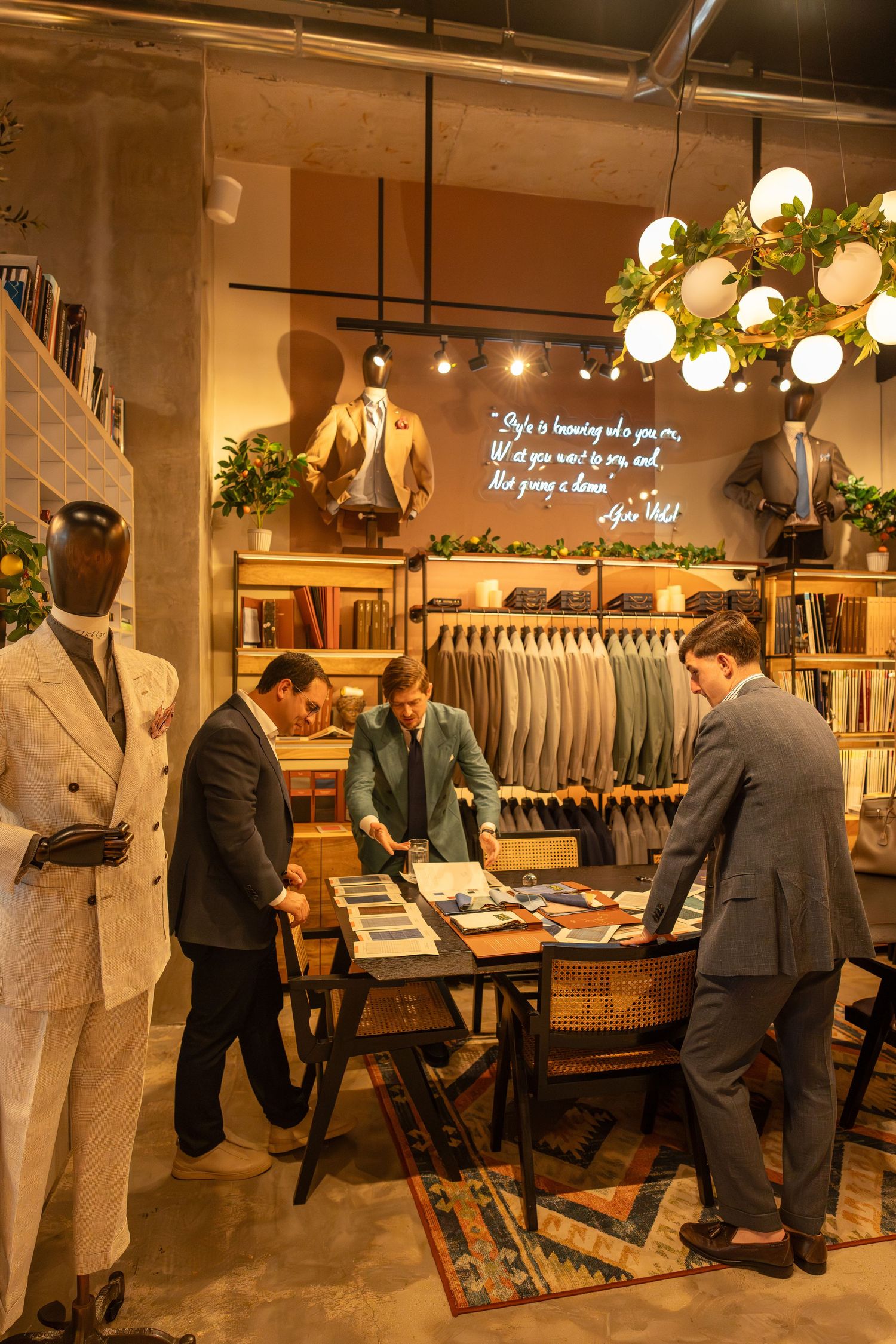 Three men in suits browse fabric samples at a table in an elegant menswear tailor shop.