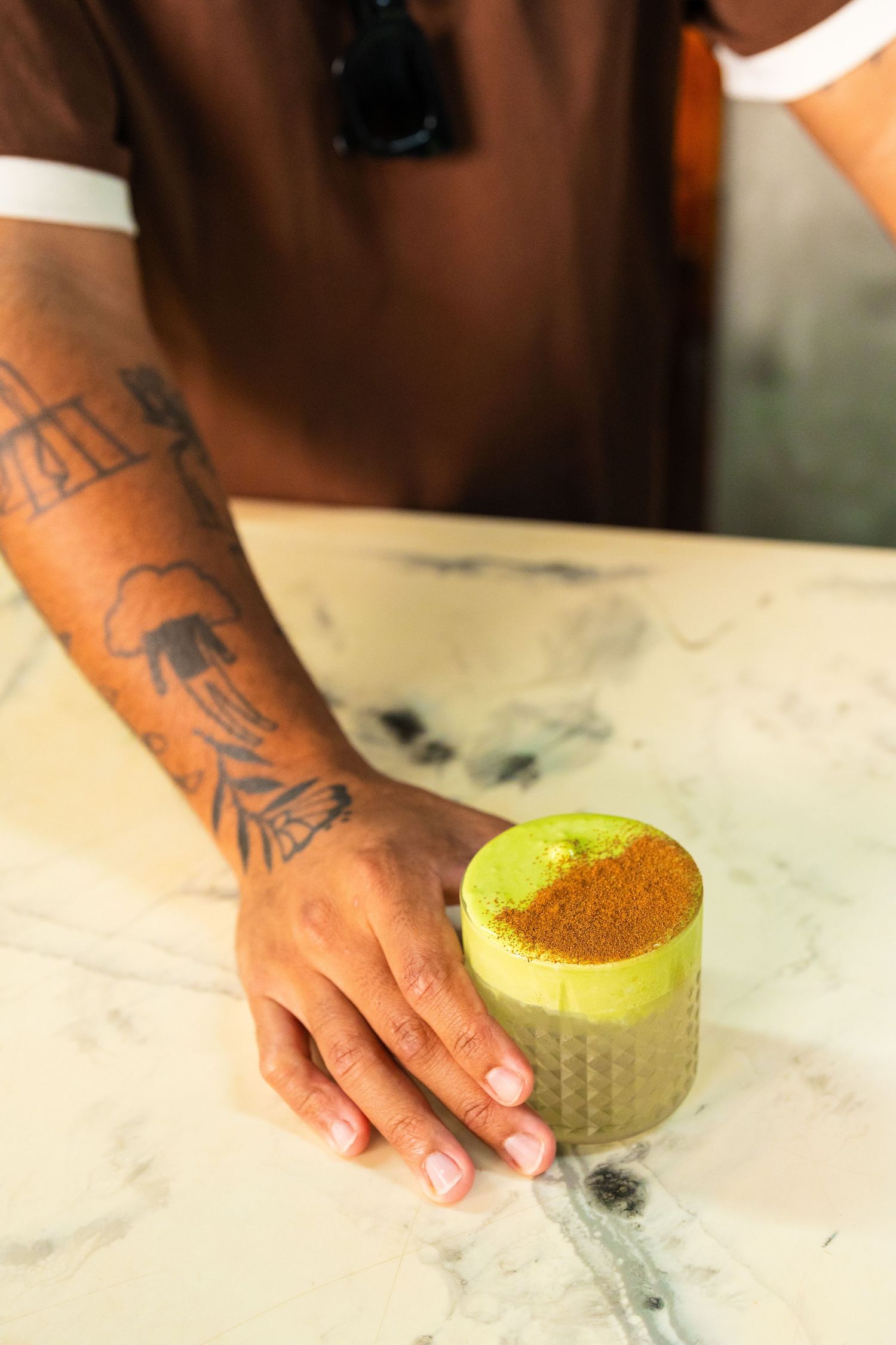 Tattooed hand resting beside a vibrant green matcha drink topped with orange spice powder on marble surface.