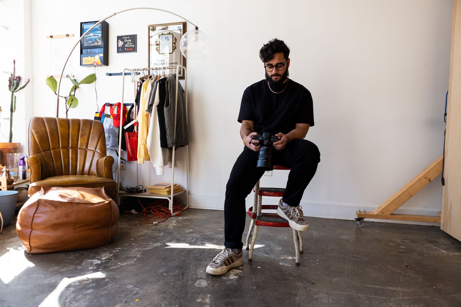 Young bearded man sitting on a stool using a camera in a bright modern studio space.
