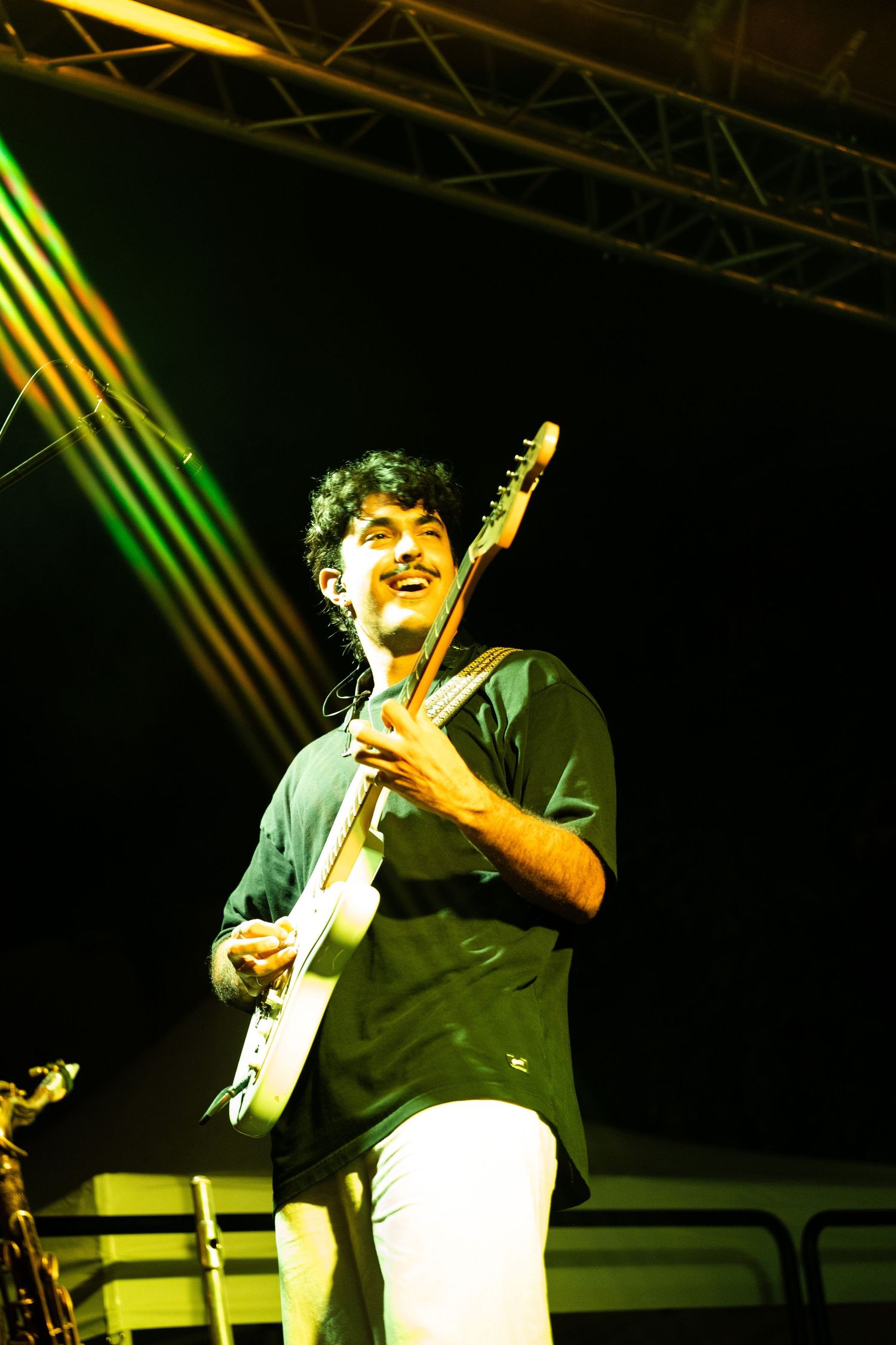 Smiling guitarist performing on stage under colorful green and yellow lights at night.