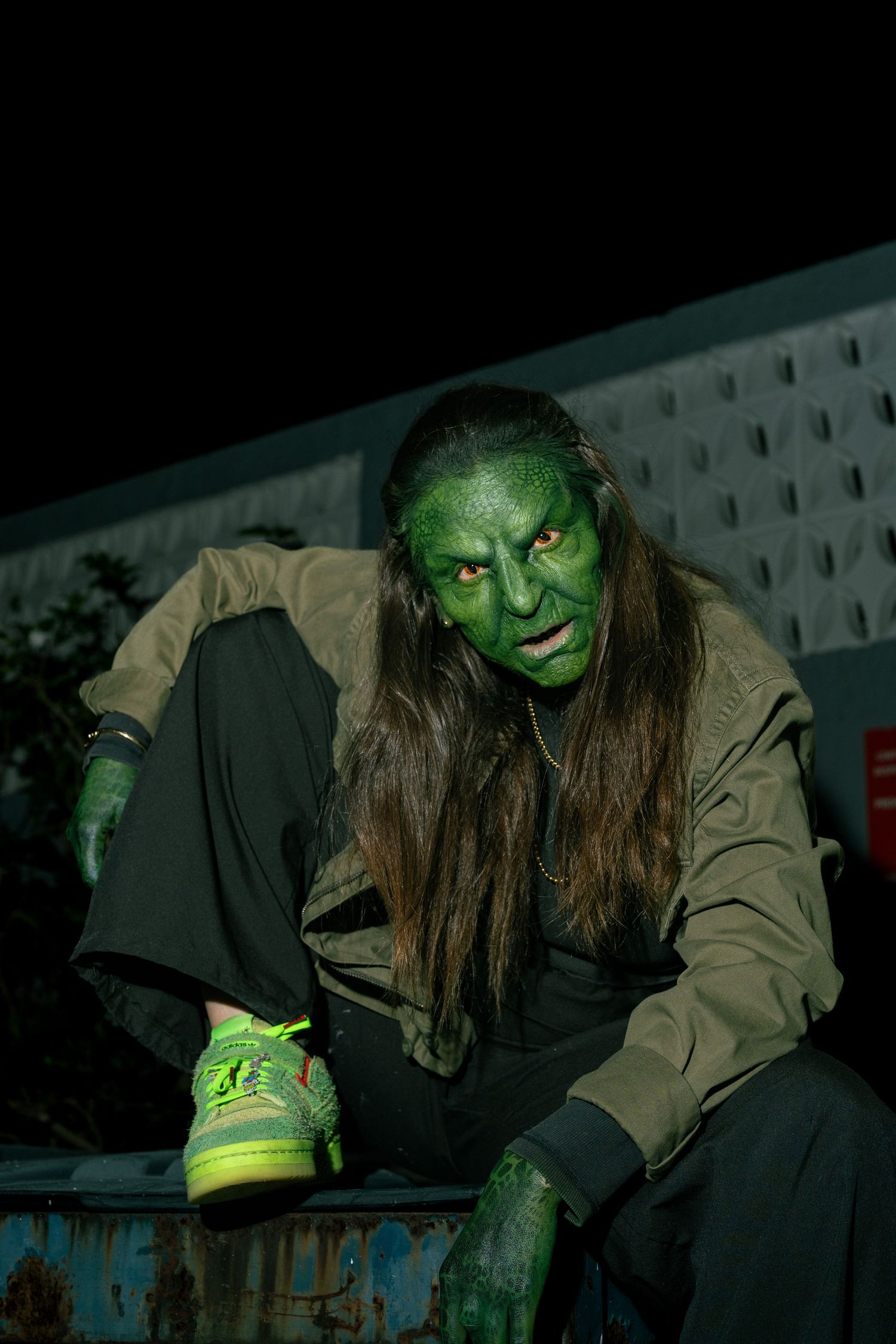 Person in green creature Halloween makeup and costume sitting on rusty surface at night.