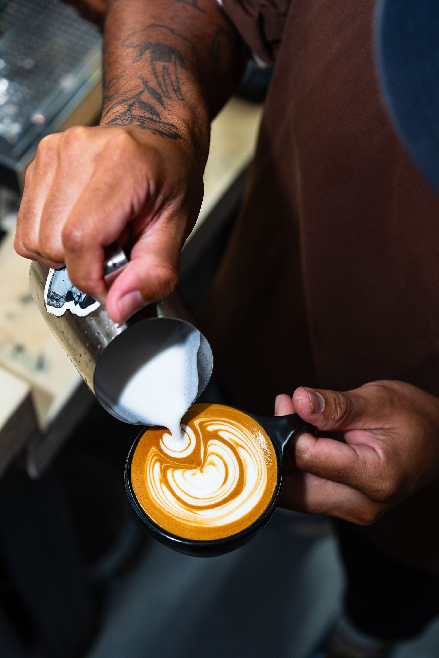Tattooed barista pouring steamed milk into a black cup creating latte art with a heart pattern design.