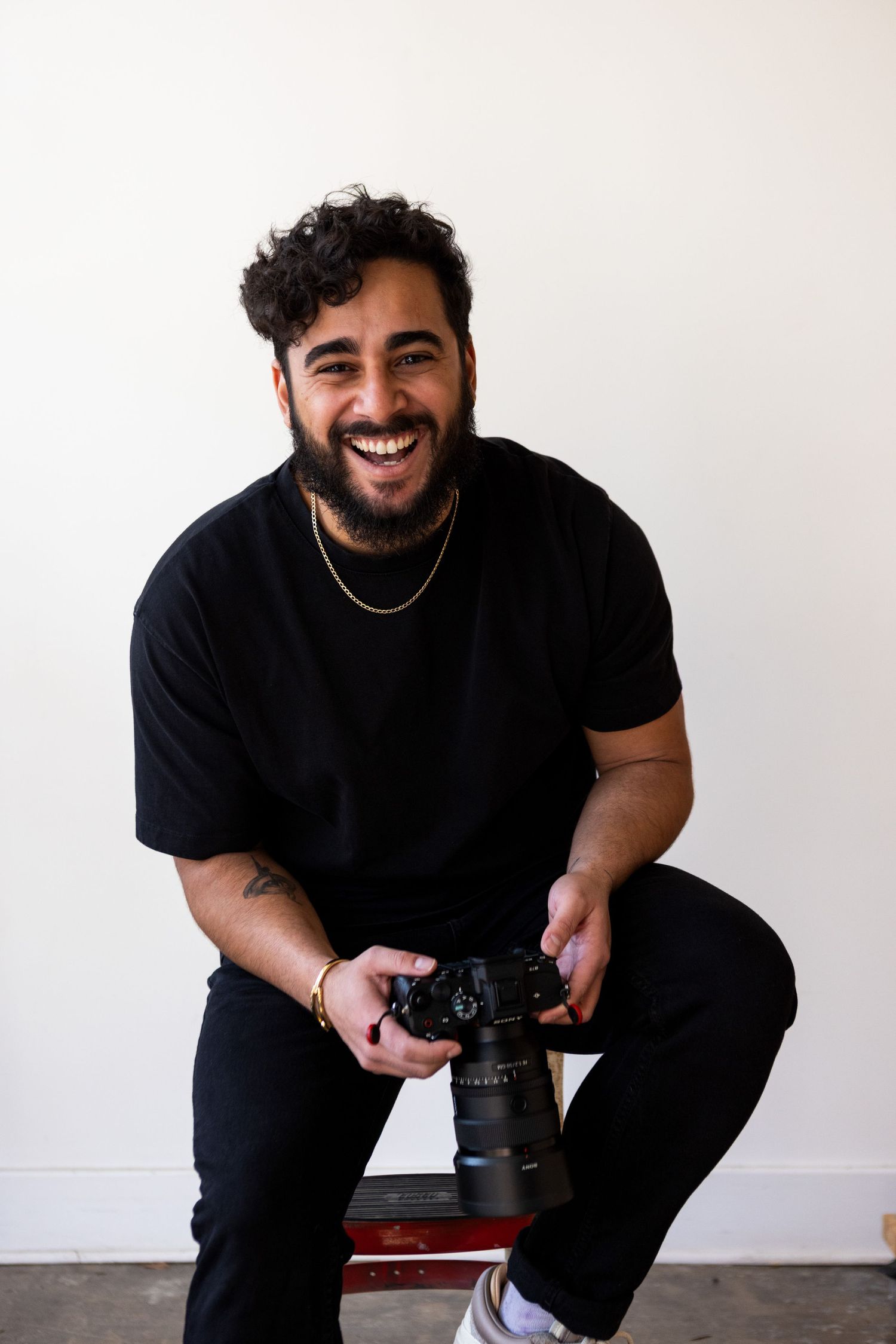Happy bearded photographer smiling while holding a professional DSLR camera against a white wall.