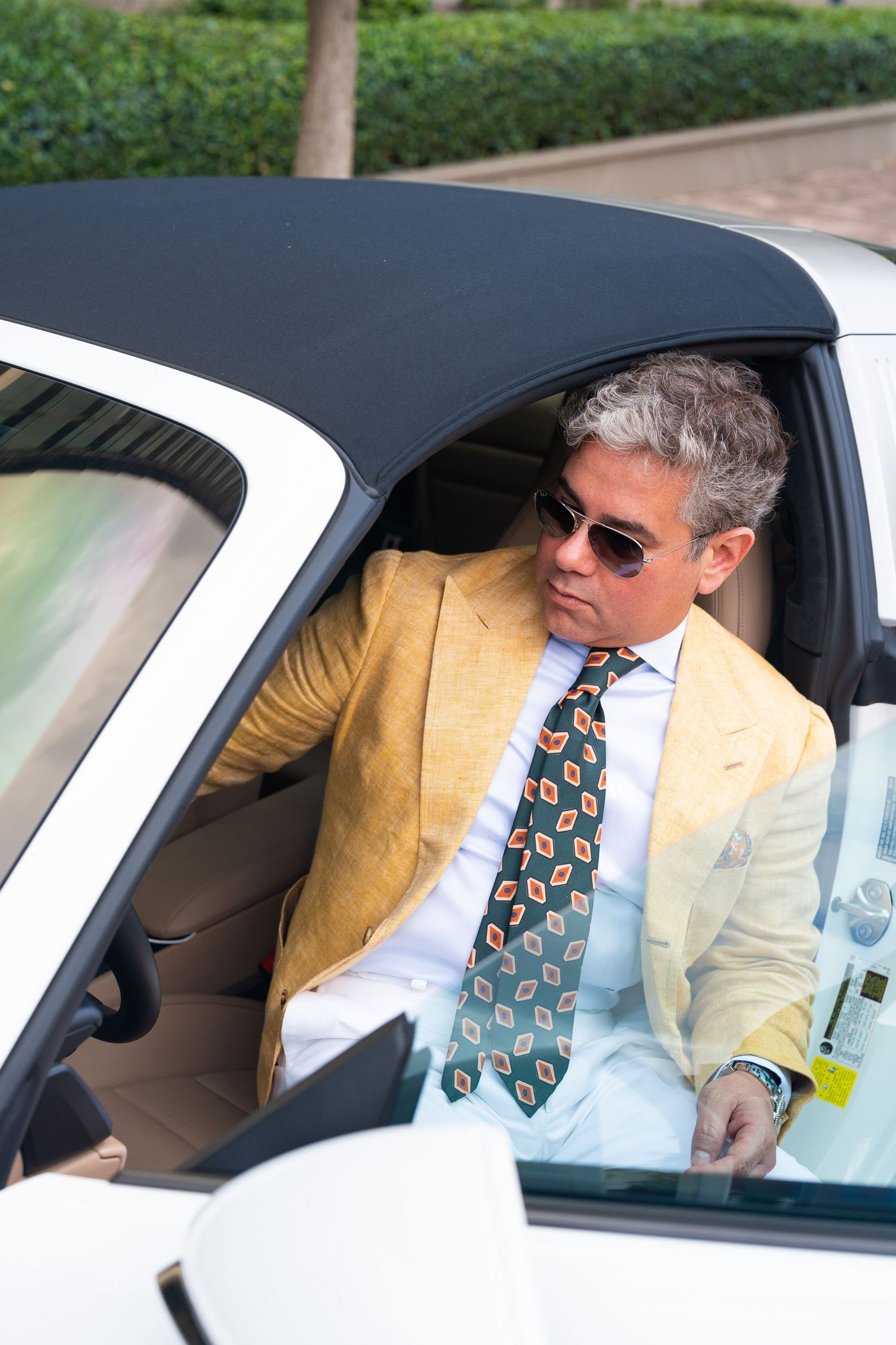 Stylish man in yellow blazer and patterned tie sitting in a white convertible car.