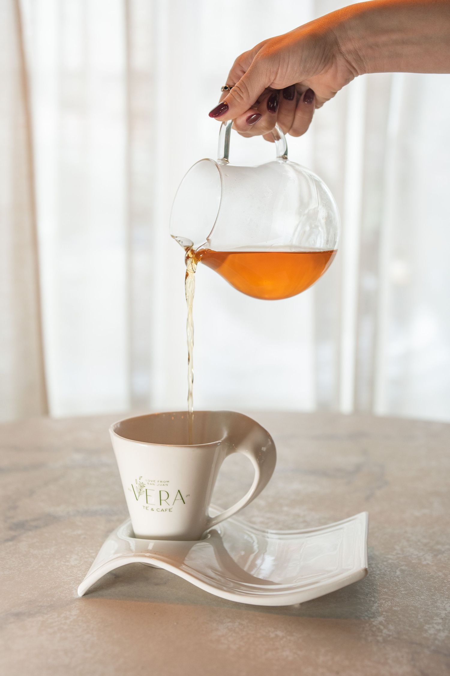 Hand pouring amber tea from glass teapot into branded Vera cafe cup on elegant white saucer.