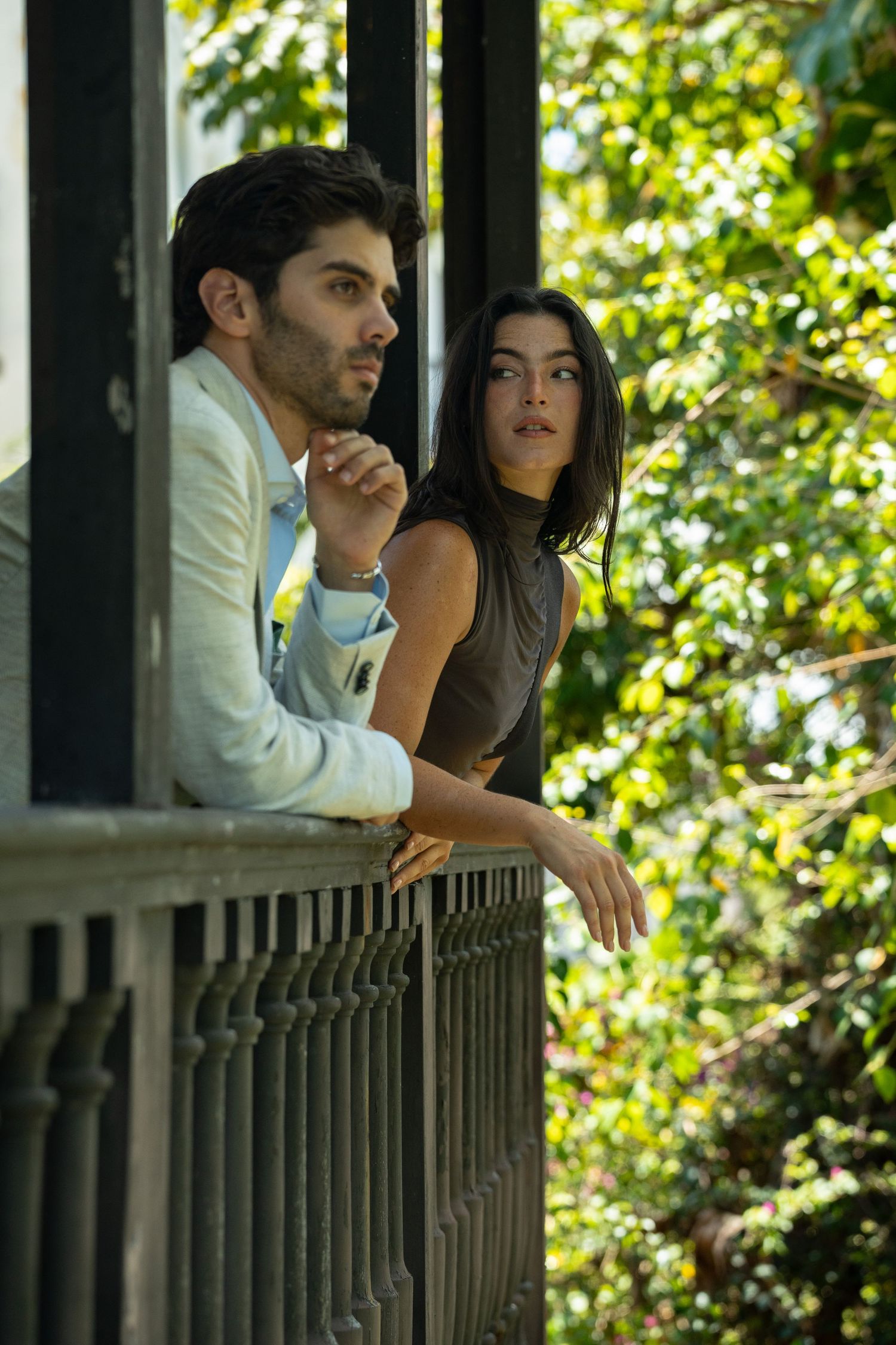 Stylish couple leaning on iron balcony railing surrounded by lush green foliage.