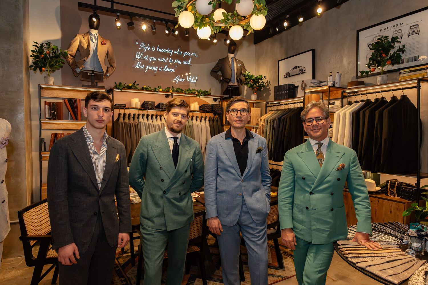 Four men in stylish suits posing inside a modern menswear clothing boutique with green and gray suit displays.