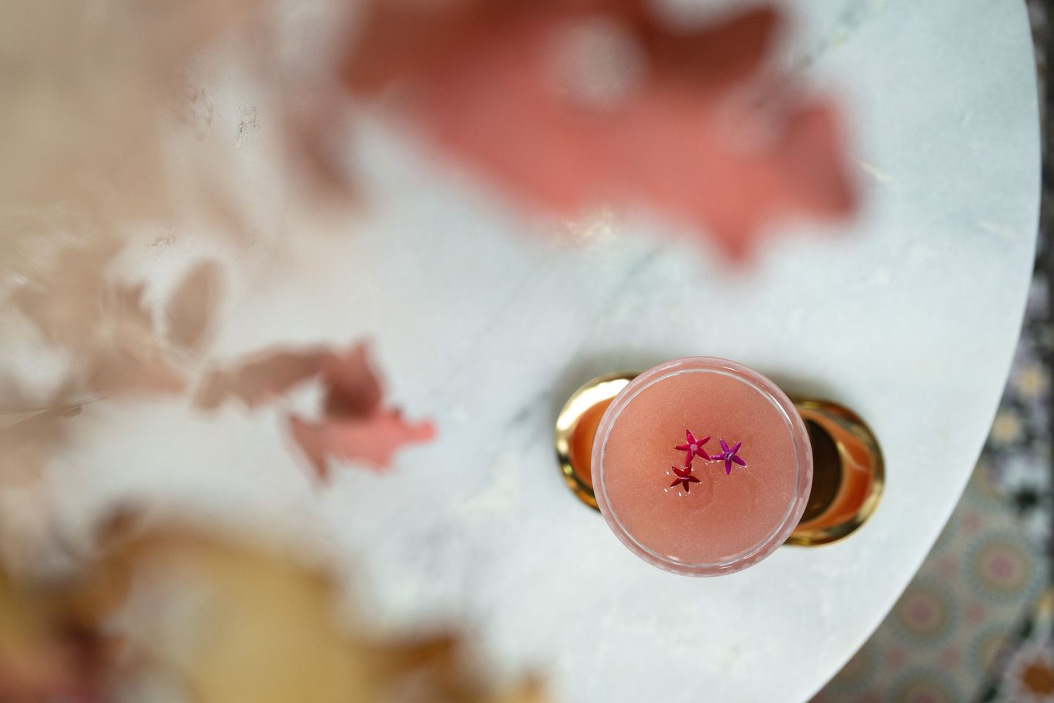 Aerial view of a blush pink cocktail with purple flower garnish on a gold coaster atop a marble table.