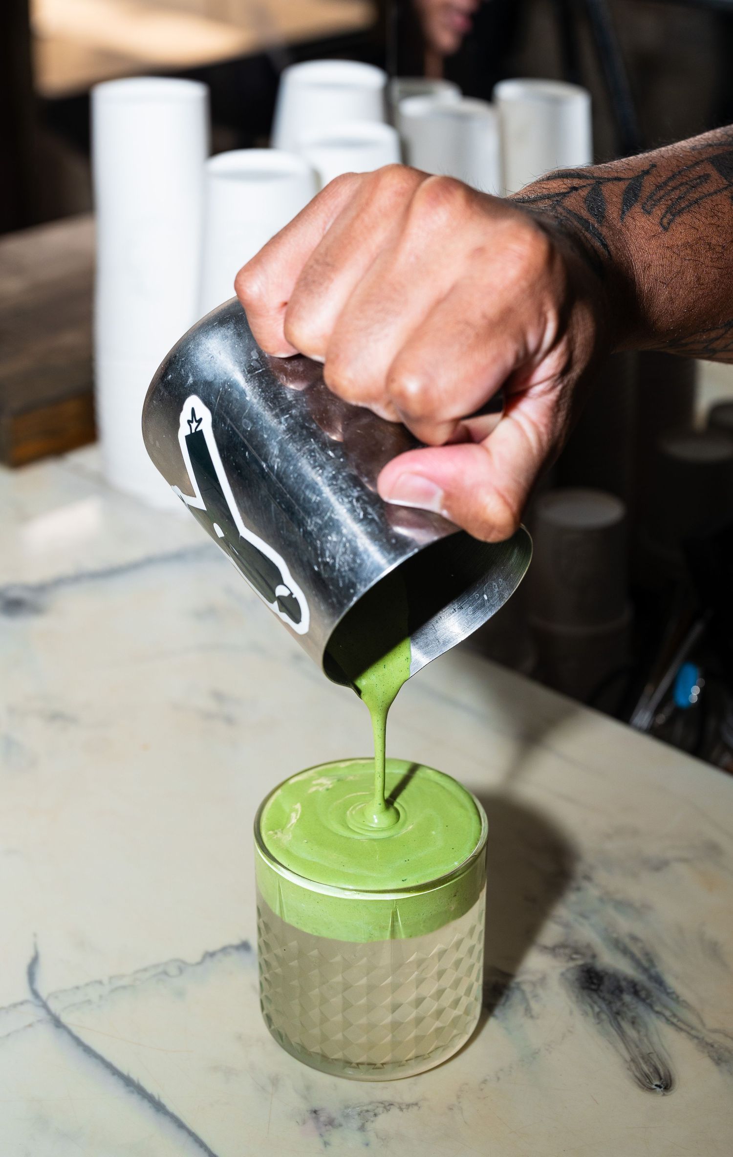 Tattooed barista pouring vibrant green matcha latte into a textured glass on a marble counter.