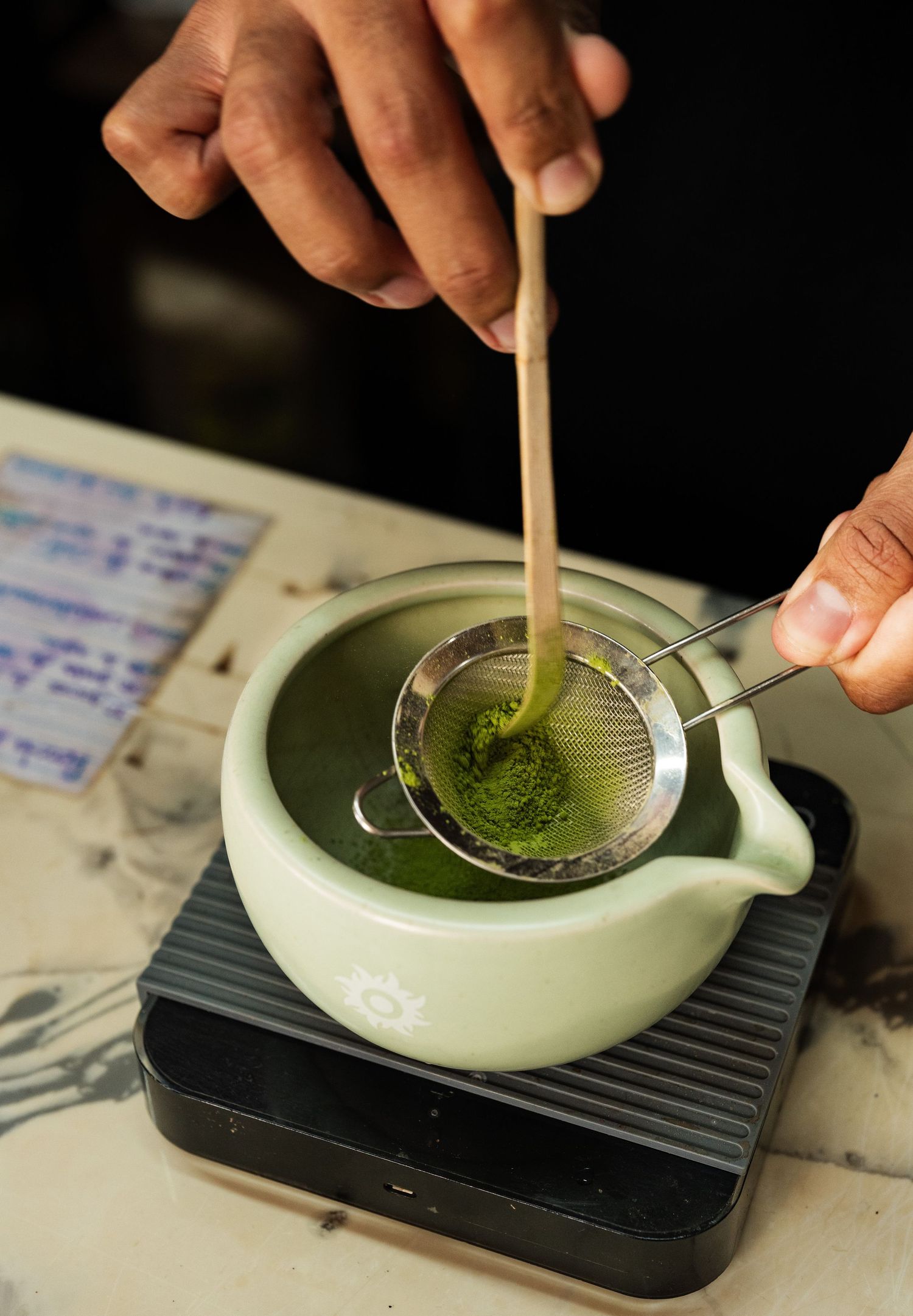 Hands sifting vibrant green matcha powder through a fine mesh strainer into a ceramic pouring bowl.