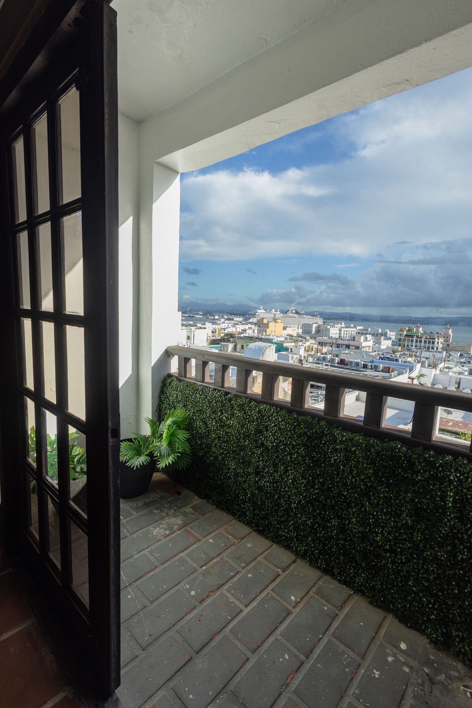 Balcony with black French doors, green hedge wall, and panoramic coastal city view.