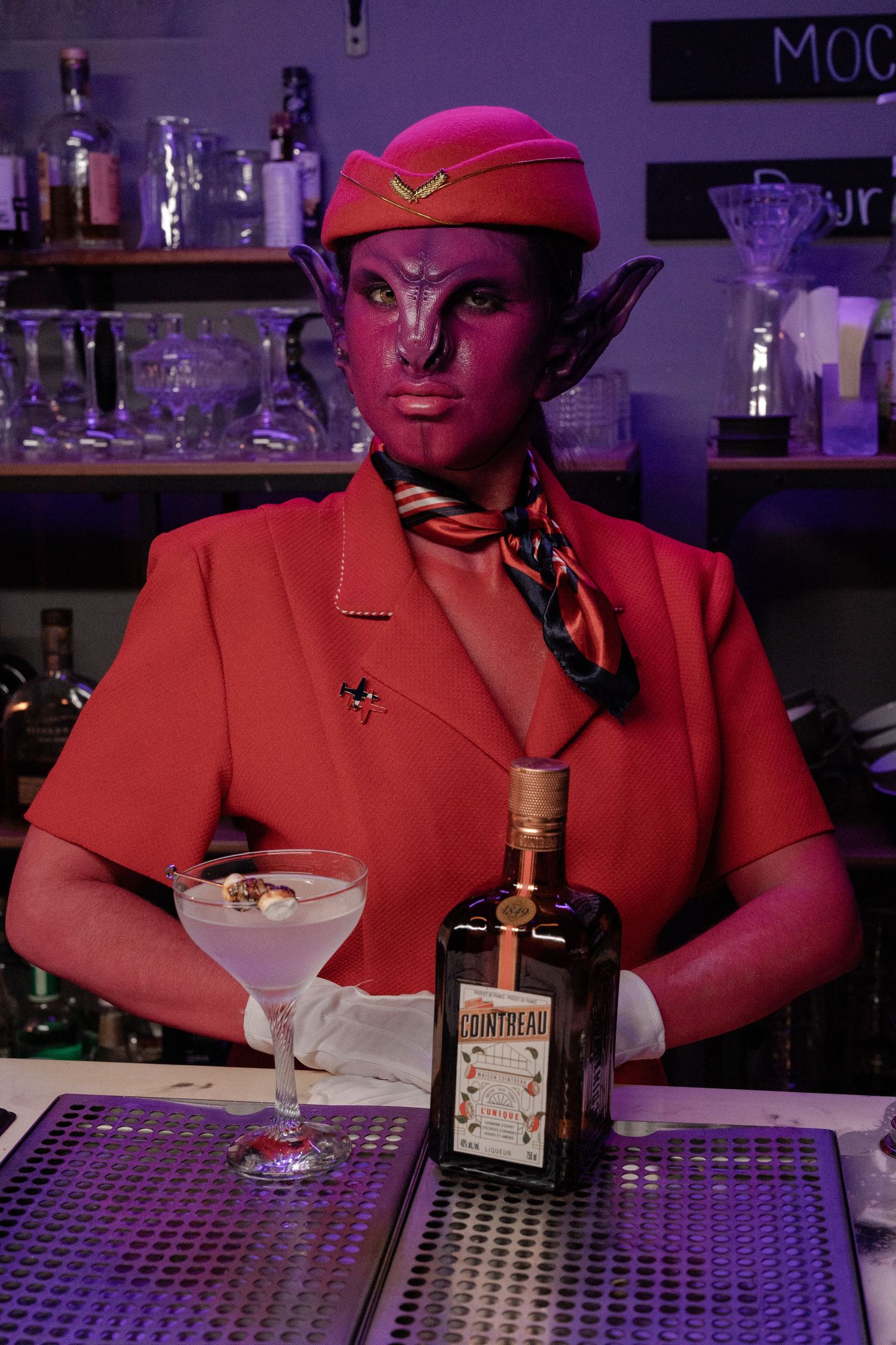 Person in purple alien makeup and red flight attendant costume posing with Cointreau bottle at bar.