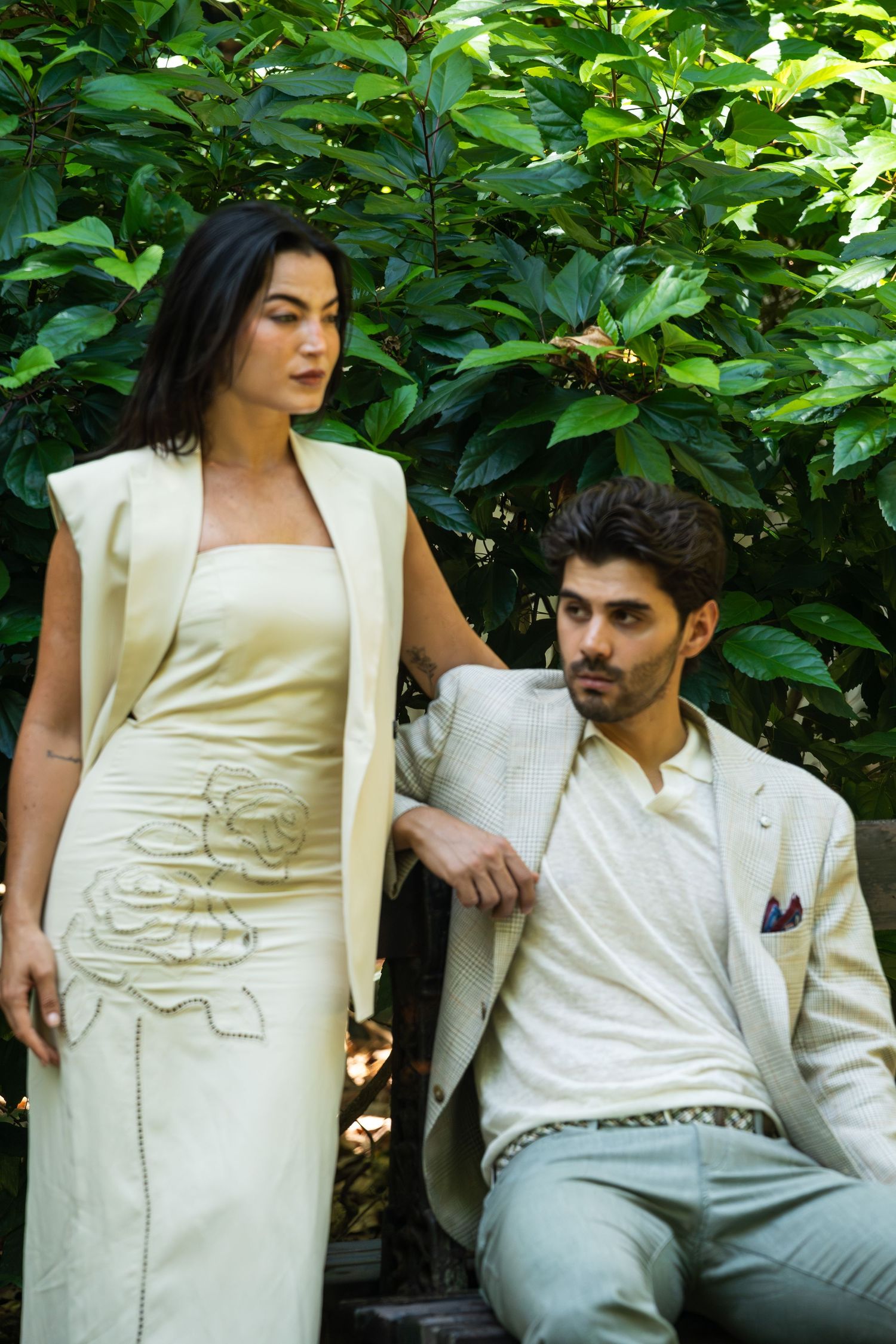 Fashion couple in cream coordinated outfits posed against lush green foliage.