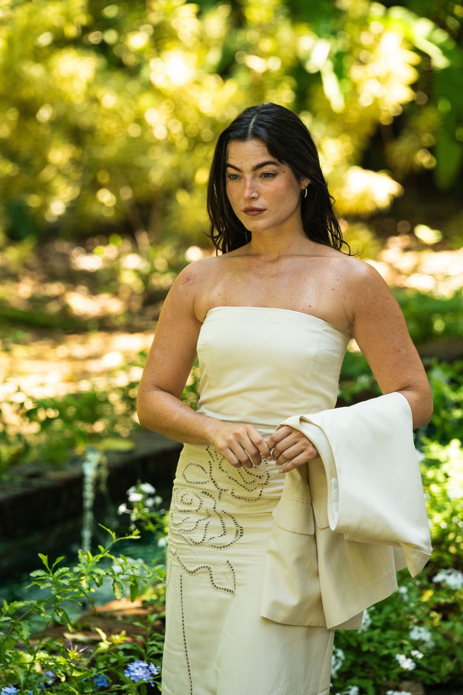 Elegant woman in cream strapless gown standing among lush green garden foliage in natural light.