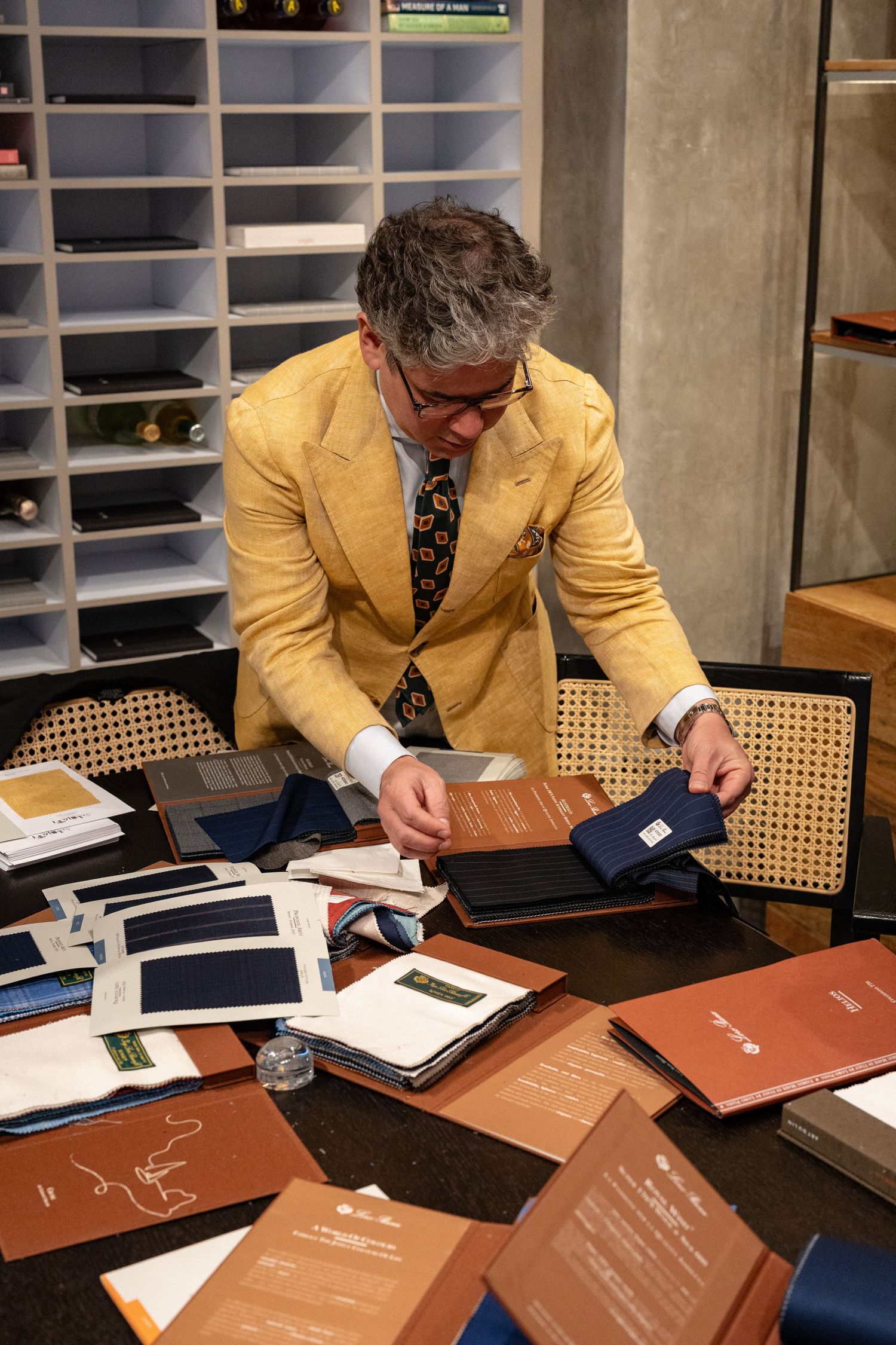 Elegantly dressed man in yellow blazer examining fabric swatches at a tailor consultation table.
