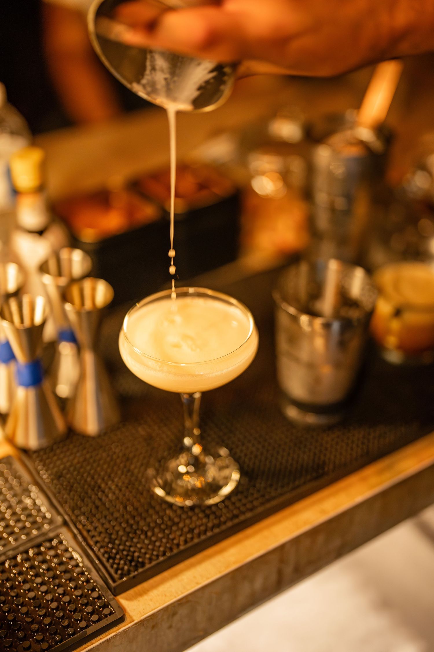 Bartender pouring creamy liquid into a coupe glass filled with a frothy pale cocktail on a bar mat.