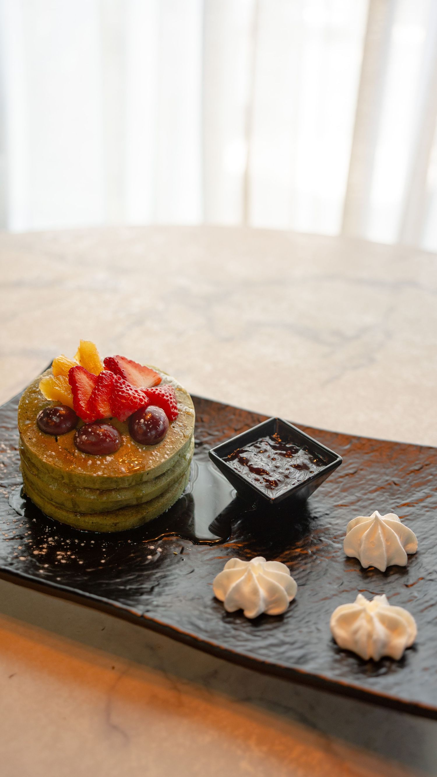 Stack of green matcha pancakes topped with fresh fruit on a dark plate with meringues and sauce.