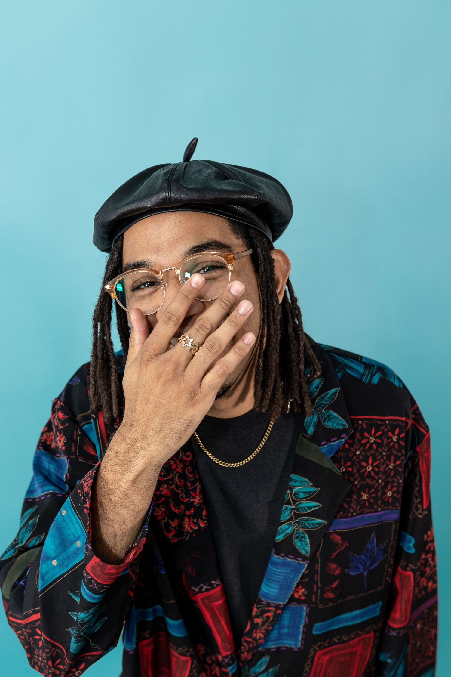 Stylish man with dreadlocks wearing beret and patterned blazer laughs against teal background.