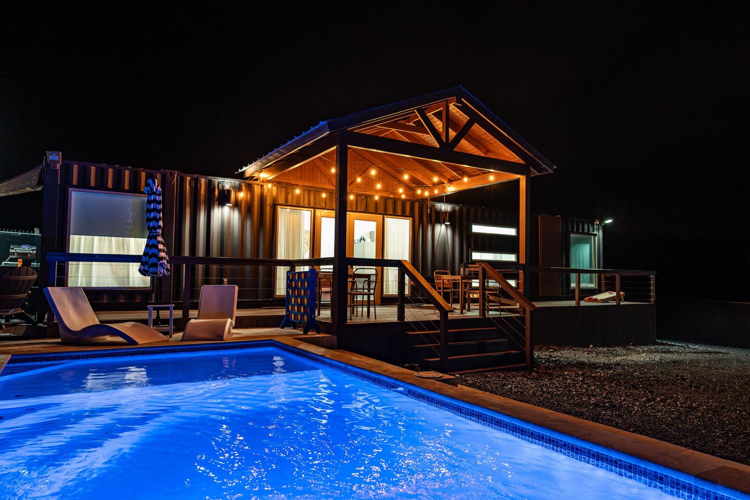 Luxury cabin with glowing string lights and a illuminated blue swimming pool at night.