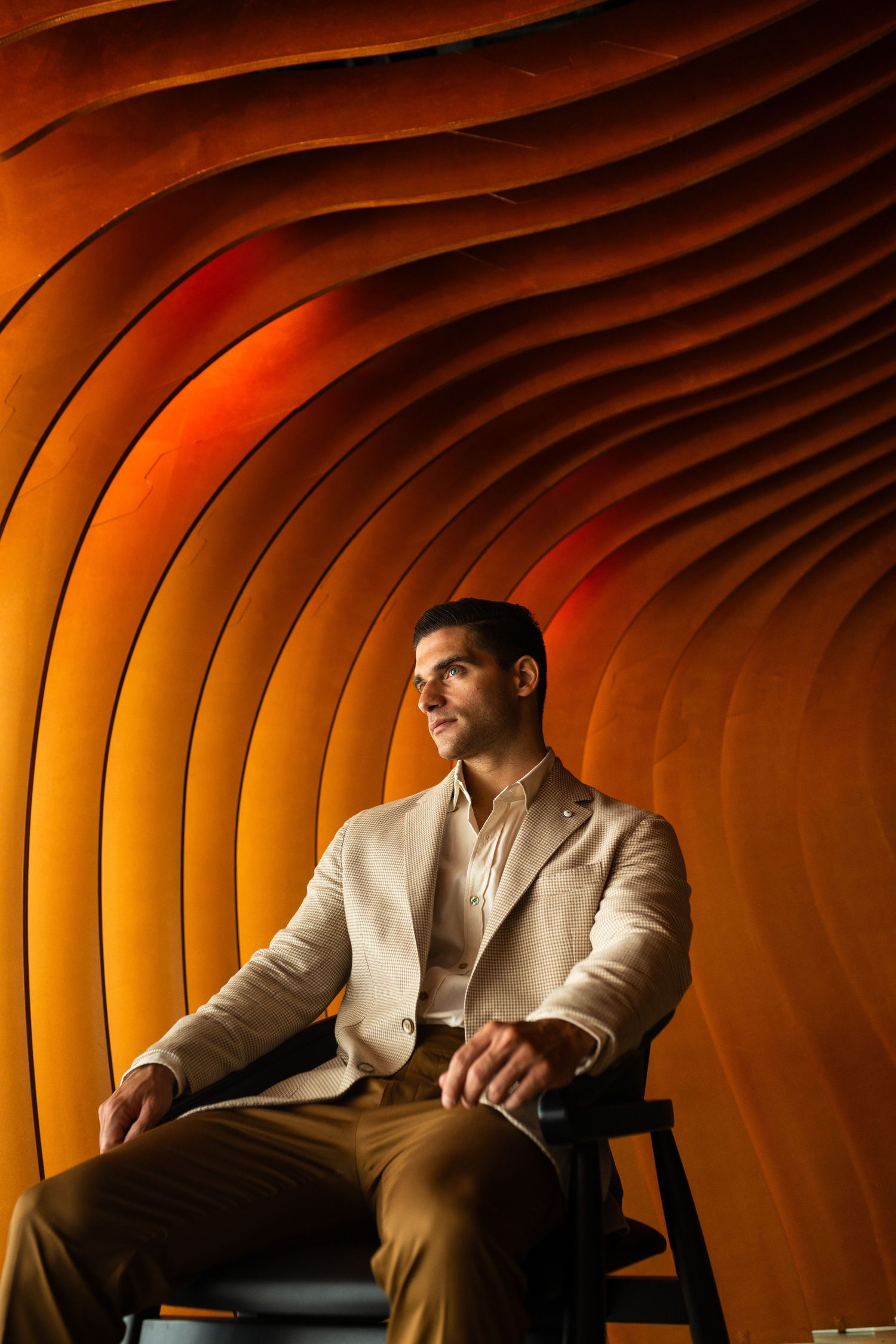 Stylish man in beige blazer sits relaxed inside a dramatic orange curved architectural tunnel.