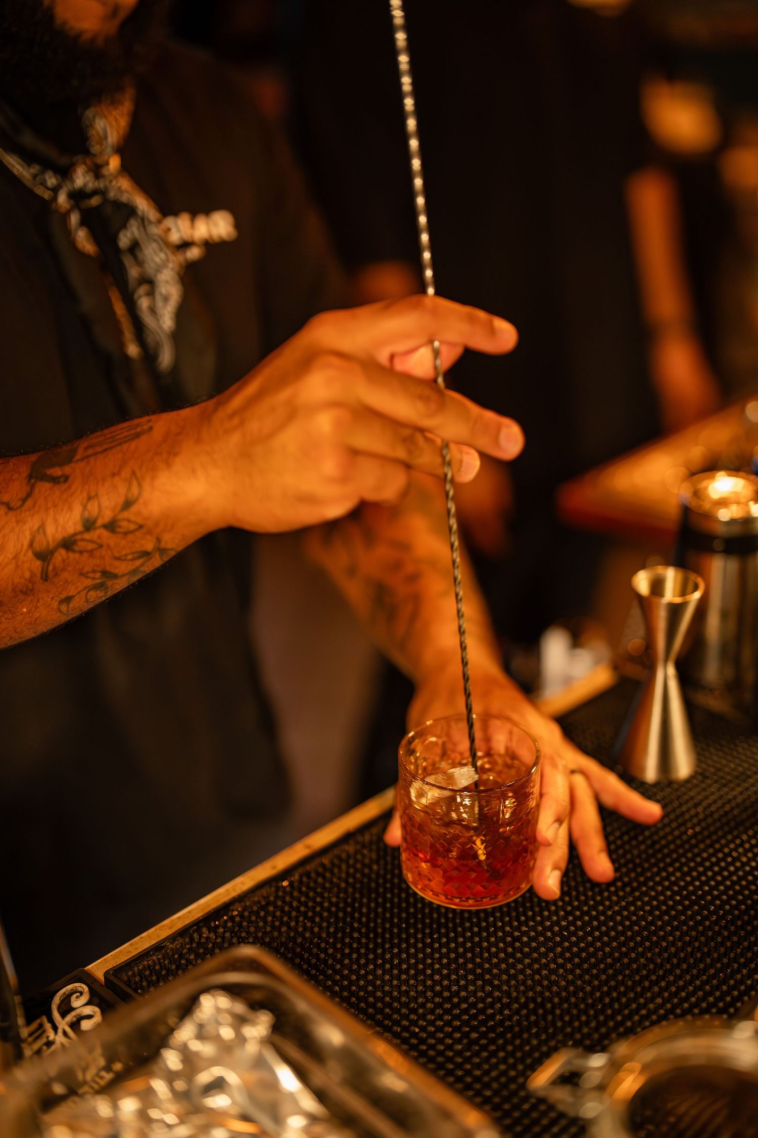 Tattooed bartender stirring a red cocktail in a rocks glass with a long bar spoon at a dimly lit bar.